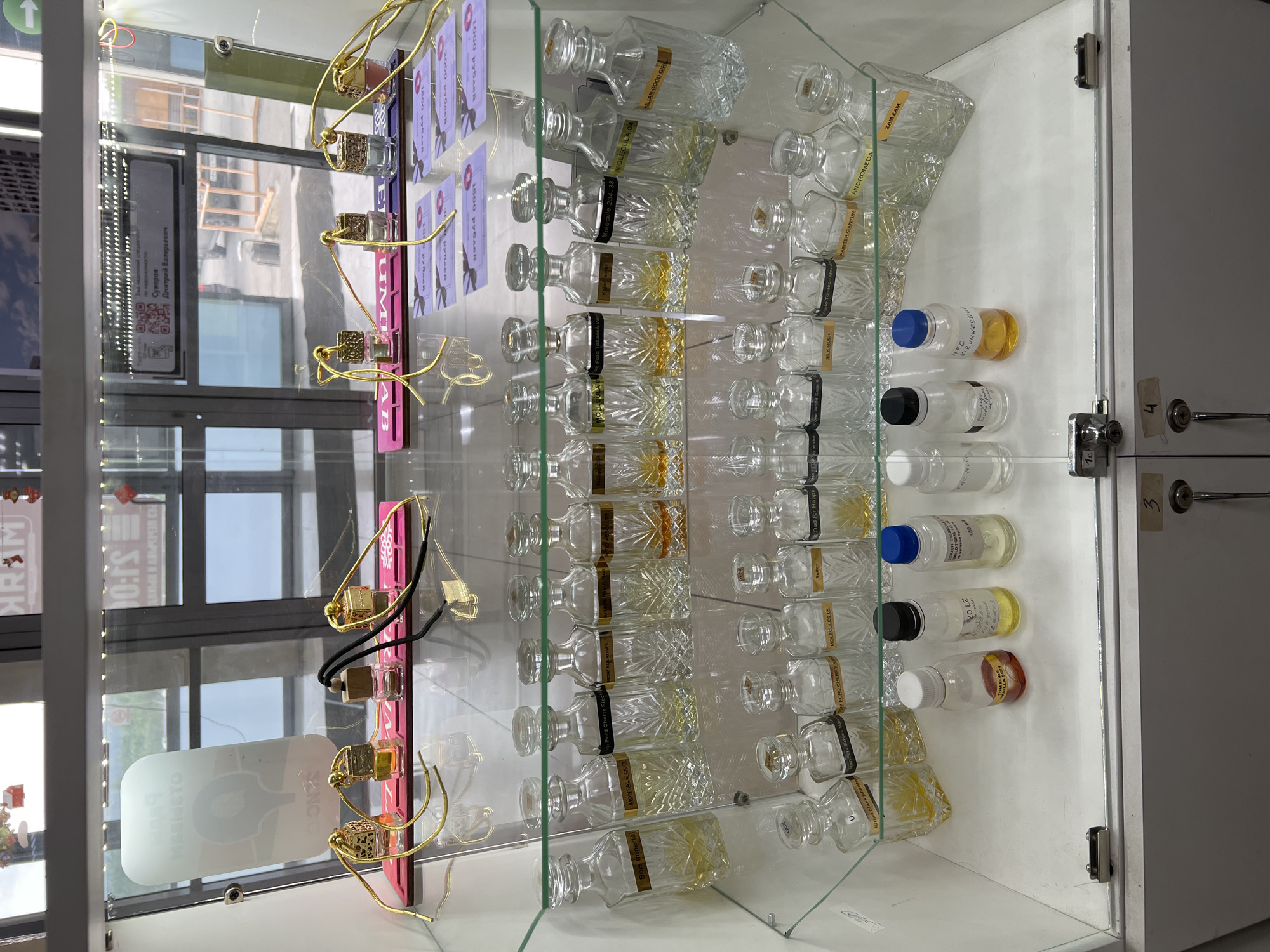This image shows a display case at a perfume shop in Yekaterinburg, Russia. The display case is made of glass and metal, and it is filled with various perfumes. In the foreground, there are several small bottles of perfume, some with colorful liquids and others empty. Behind these, there are rows of larger, ornate glass bottles, many of which appear to be full of perfume. Some of the bottles have labels indicating brand names or scents. The top shelf of the display case features a collection of small, square-shaped perfume bottles with gold accents and decorative hanging elements. In the background, through the glass of the display case, reflections of the store's interior and the street outside are visible, including shop signage and windows. The overall impression is of a well-stocked perfume store showcasing a variety of fragrances.