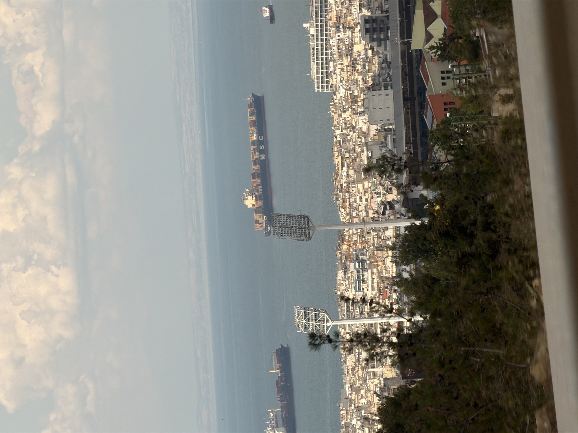 The image captures a wide-angle, elevated view of the city of Thessaloniki, Greece, overlooking the Thermaic Gulf. The foreground is dominated by dense green foliage, suggesting the viewpoint is from a hillside or a high vantage point within the city. Partially obscured by the trees are two tall, white stadium floodlights, one of which is more prominently featured on the left side of the frame.
Beyond the floodlights, the urban landscape of Thessaloniki unfolds. Buildings with light-colored facades, many appearing to be multi-story apartments or commercial structures, densely pack the city. Some buildings exhibit balconies and varied architectural details, indicating a mature urban environment. A large, modern white building with numerous windows stands out in the middle ground.
The Thermaic Gulf occupies the background, its waters a deep blue under a sky filled with a mix of bright white and softer gray clouds. Two large cargo ships are visible on the water. The most prominent one, a container ship, is dark-hulled with a vast deck covered in colorful shipping containers, and it bears the "MSC" logo. A smaller vessel, likely another cargo ship or a tugboat, is positioned further to the right and in the distance. The horizon line is a faint separation between the sea and a hazy landmass in the far distance. The overall lighting suggests it is daytime, possibly late afternoon or early morning, given the soft light and cloud cover. The scene depicts a bustling port city with significant maritime activity, viewed from a scenic overlook.