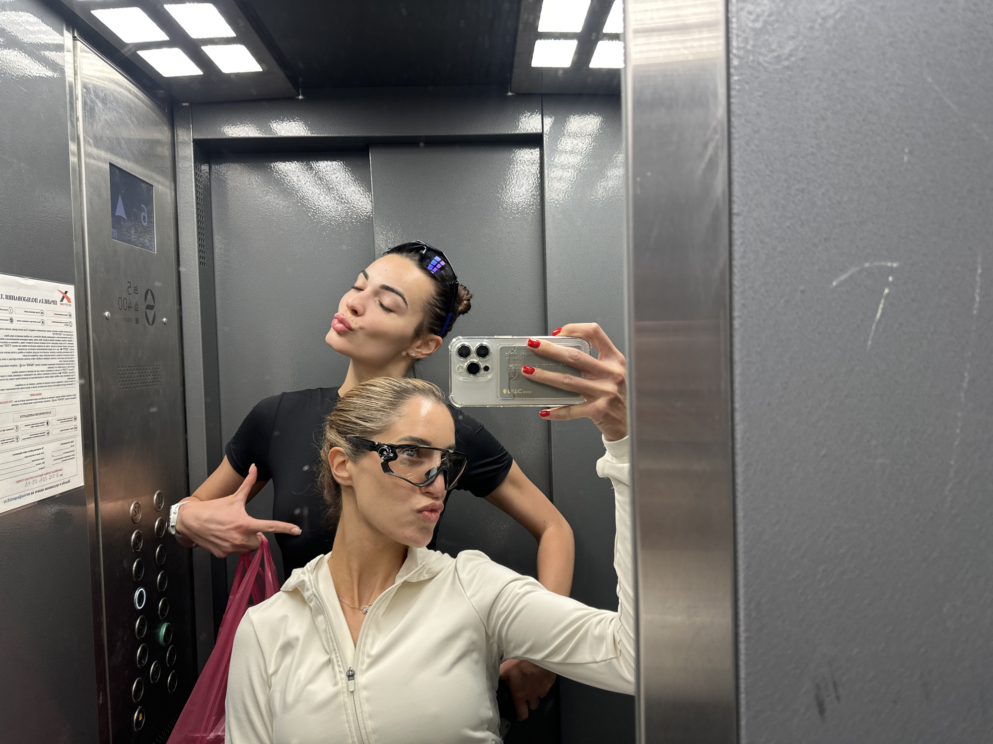 Two women are taking a selfie in an elevator. The woman in the foreground is wearing a white zip-up hoodie and black sunglasses, and has her hair pulled back. The woman behind her is wearing a black t-shirt and has her hair in a bun, and is making a peace sign with her fingers. There is a pink bag in the background. The elevator appears to be in a residential building. The overall scene is candid and informal. The location is not identifiable as Moscow, Russia.