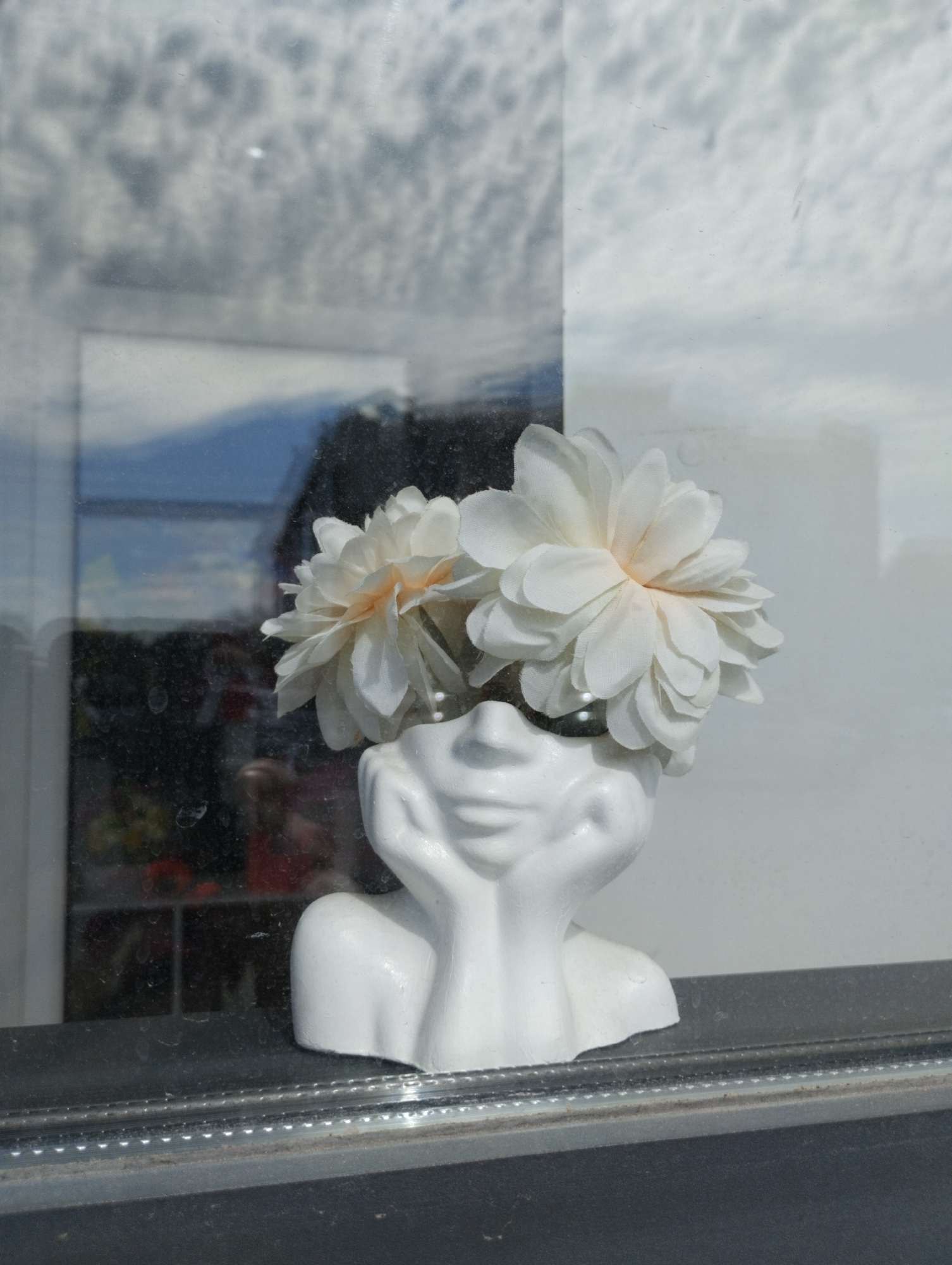 The image shows a white ceramic vase in the shape of a human bust, placed on a windowsill. The vase is adorned with two white fabric flowers, positioned as if they are emerging from the top of the bust's head. The bust has its hands resting on its cheeks, in a contemplative or resting pose. The vase is seen through a glass window, which reflects the sky and some blurred background elements. In the background, through the window, there is a reflection of a cloudy sky and a suggestion of a building or an interior space with some indistinct colored objects. The windowsill appears to be made of dark material, with some visible dirt or dust. The lower part of the image shows the edge of the windowsill and a metallic strip with some small lights. The scene appears to be an indoor display, possibly in a shop or a home, with the window providing a view of the outdoors. The location is identified as Kostanay, Kazakhstan.
