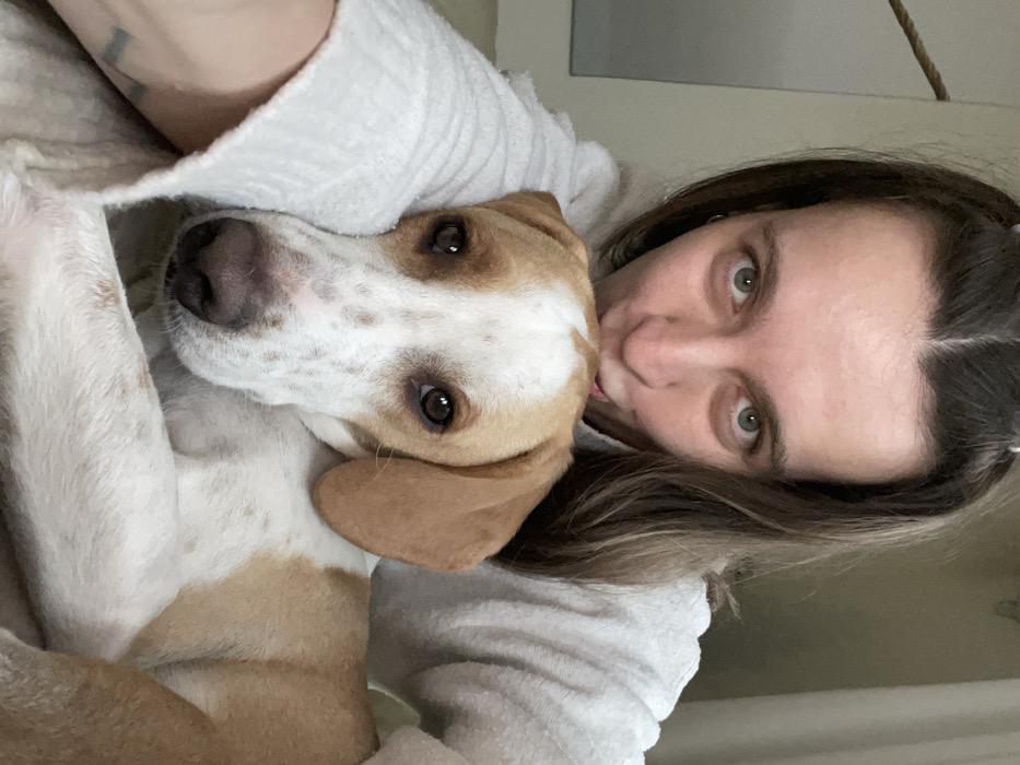A close-up indoor portrait features an adult female and a dog. The female, positioned at the top of the frame, has long, dark-brown hair with a center part, light-colored eyes, and a light complexion. She wears a light-colored, textured sweater, possibly white or light grey. Her face is tilted, and her mouth is positioned over the dog's head, obscured from view. A thin silver hoop earring is visible in her left ear, and a faint tattoo is present on her left forearm. The dog, a medium-sized canine, occupies the lower portion of the frame. It has predominantly white fur with tan/brown patches on its head, ears, and parts of its body, and brown speckled markings on its white snout. The dog has dark brown eyes and floppy ears. It is resting its head against the female's body or arm. Both subjects are looking directly into the camera. The background is blurred, depicting neutral-toned surfaces and a rope-like object in the upper left. The specific location within the city of Budva, Montenegro, cannot be determined from the visual evidence.