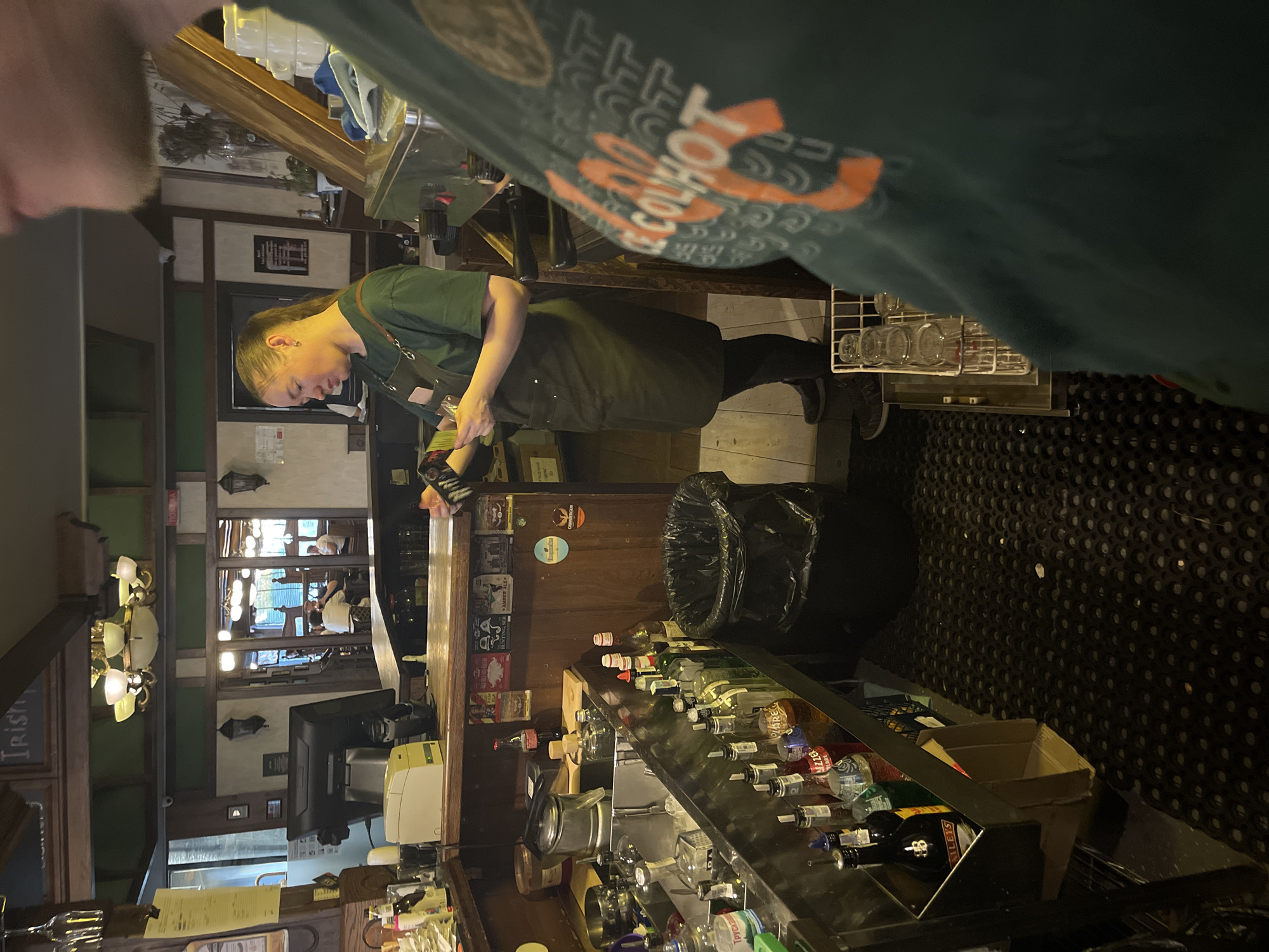 The image shows a bar area, likely in a pub or restaurant, with a bartender working behind the counter. The bartender, a woman with her hair tied back in a ponytail, is wearing a dark apron over a green t-shirt and is holding a small device, possibly a card reader or scanner. In the foreground, part of a person wearing a green t-shirt with orange lettering is visible on the right side of the frame. The bar itself is stocked with numerous bottles of alcohol and other bar supplies. There is a sink area with ice and bottles, and a trash can is located to the right of the sink. In the background, through an open doorway, other patrons can be seen seated at tables in a dimly lit dining area. The overall decor suggests an Irish pub or a similar establishment, with wooden beams and traditional lighting fixtures. The scene appears to be indoors. There is no information available to confirm the location in Kyiv, Ukraine.
