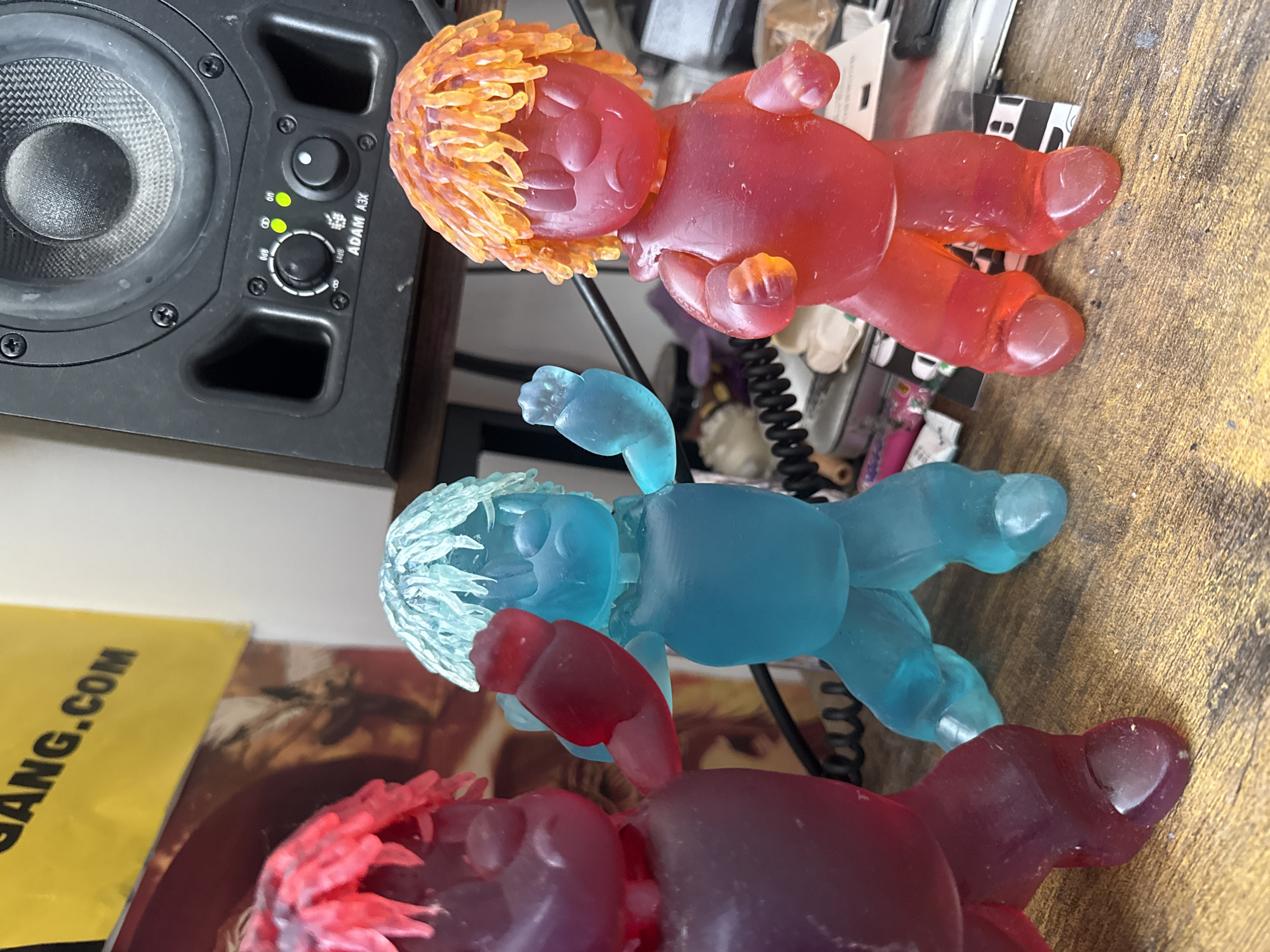 The image captures a rotated view of three translucent, resin-like figurines resembling stylized human figures, displayed on a wooden surface next to a studio monitor speaker.
The three figurines vary in color and are positioned in a line. The foreground figure, partially visible, is dark red or purple with red, textured hair. The middle figure is bright blue with light blue, textured hair, and the background figure is bright red with fiery orange, textured hair. All figures have simplified features, including closed eyes and a gummy or jelly-like appearance due to their translucent material.
To the left, a black ADAM A3X studio monitor speaker is visible. Its main speaker cone features a woven texture, and below it are control knobs and two illuminated green indicator lights. Text labels "OFF", "14dB", "50Hz", and "1kHz" are visible around the controls, along with the brand name "ADAM A3X".
The figurines and speaker rest on a textured wooden surface, possibly a desk. The background beyond the figures shows some general clutter and faint reflections. Part of a yellow object or paper with the text "GANG.COM" is visible in the lower left corner.
The entire image is rotated 90 degrees counter-clockwise. The scene appears to be indoors under ambient lighting. The provided location context is London, United Kingdom.