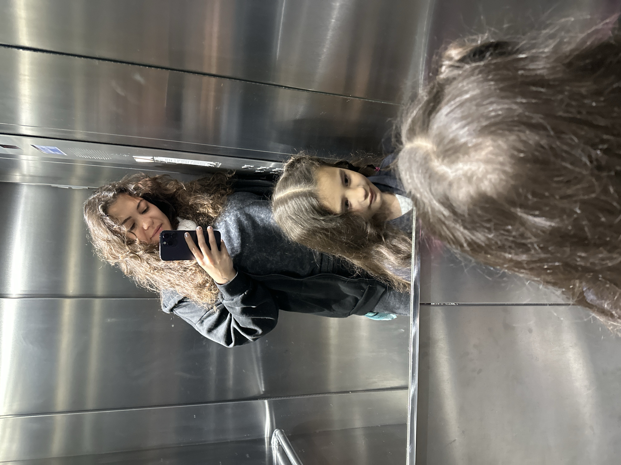 In an elevator with stainless steel walls, two females are visible. One female is holding a smartphone and taking a selfie. The reflection reveals another person present in the elevator, a child with long hair. It is not possible to determine the location as Budva, Montenegro, from the image.