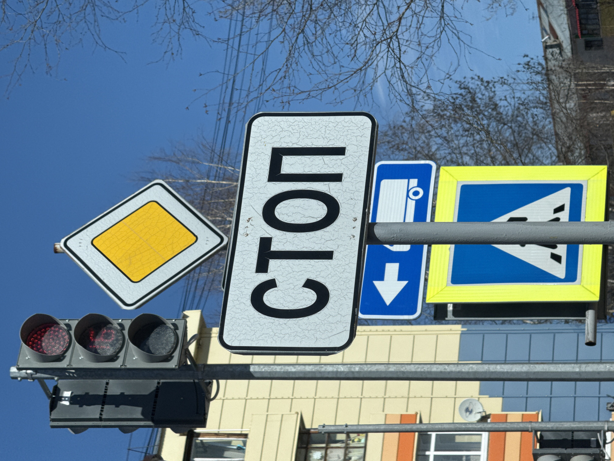 The image shows a set of traffic lights and several road signs in Beloyarsky, Russia. The traffic lights display a red light. The road signs include a priority road sign (yellow diamond), a "СТОП" (STOP) sign in Cyrillic, a bus stop sign, and a give way sign. Bare tree branches and a bright blue sky are visible in the background. The structures of buildings are also present in the background.