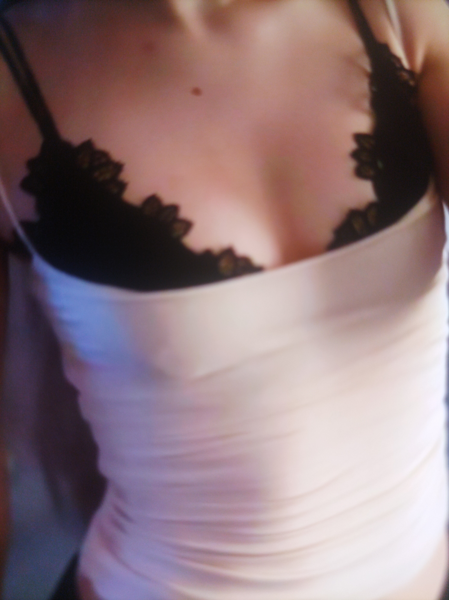 The image shows a close-up, blurry view of a person's upper torso. They are wearing a light-colored, possibly pink or beige, ribbed tank top over a black bra with lace detailing. The background is dark and out of focus. There are no discernible details about the location or any specific actions taking place.