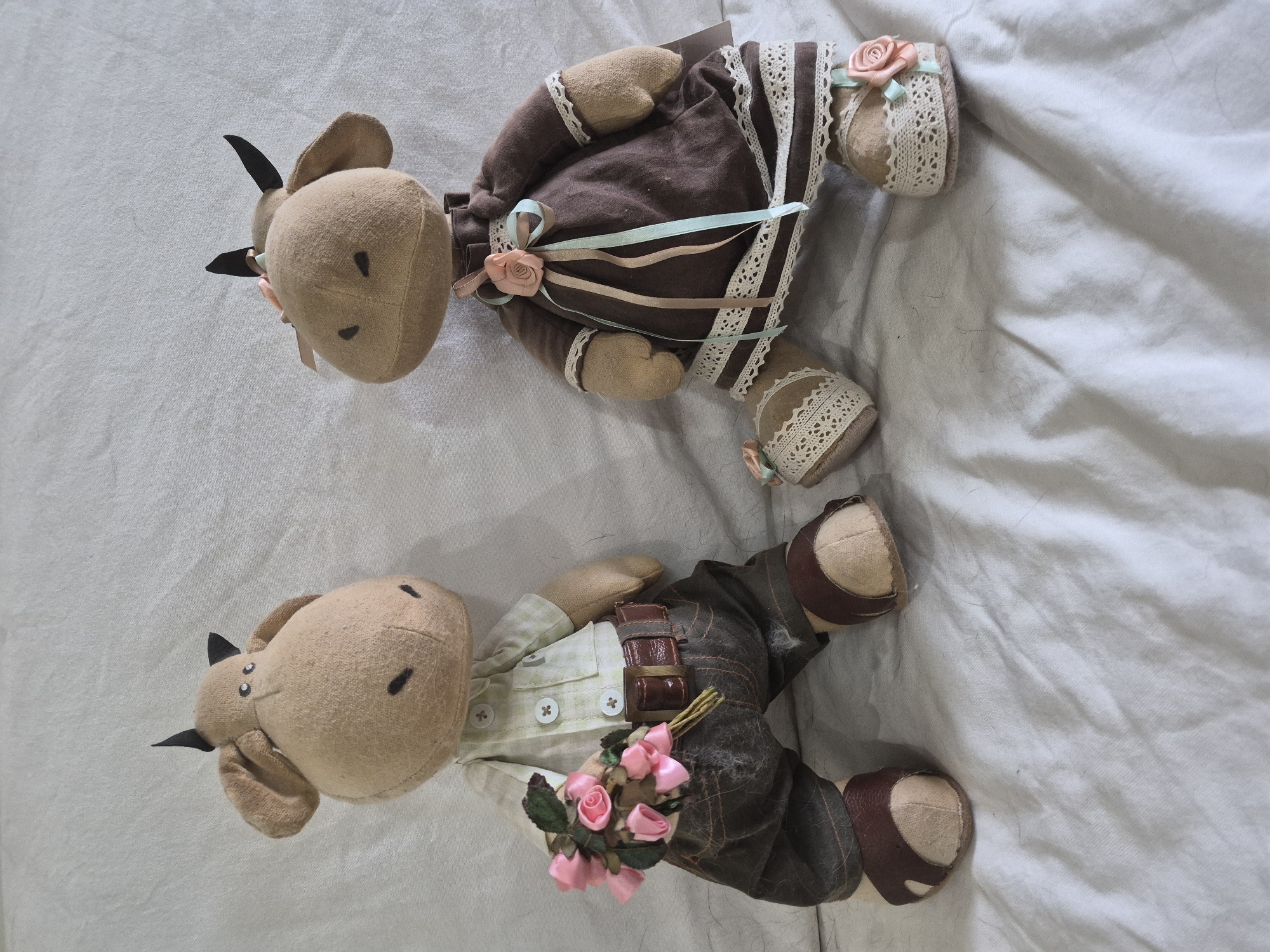 This is a medium shot, eye-level, indoor photograph of two stuffed cow figures sitting side-by-side on a white, creased cloth surface. The figures are handmade, appearing to be crafted from a beige fabric. The figure on the left is dressed in a light green collared shirt with white buttons, dark brown pants with visible stitching, and brown sandals. It is holding a small bouquet of pink fabric roses in its left hand. The figure on the right is wearing a dark brown dress with white lace trim at the hem and cuffs. It has ribbons of light brown and light blue tied at the waist. Both figures have black triangular horns and are facing slightly towards each other, suggesting an interaction. The background is a plain, white, creased cloth. There is no information in the image to suggest a specific location in the city of Vlast Truda, Russia.