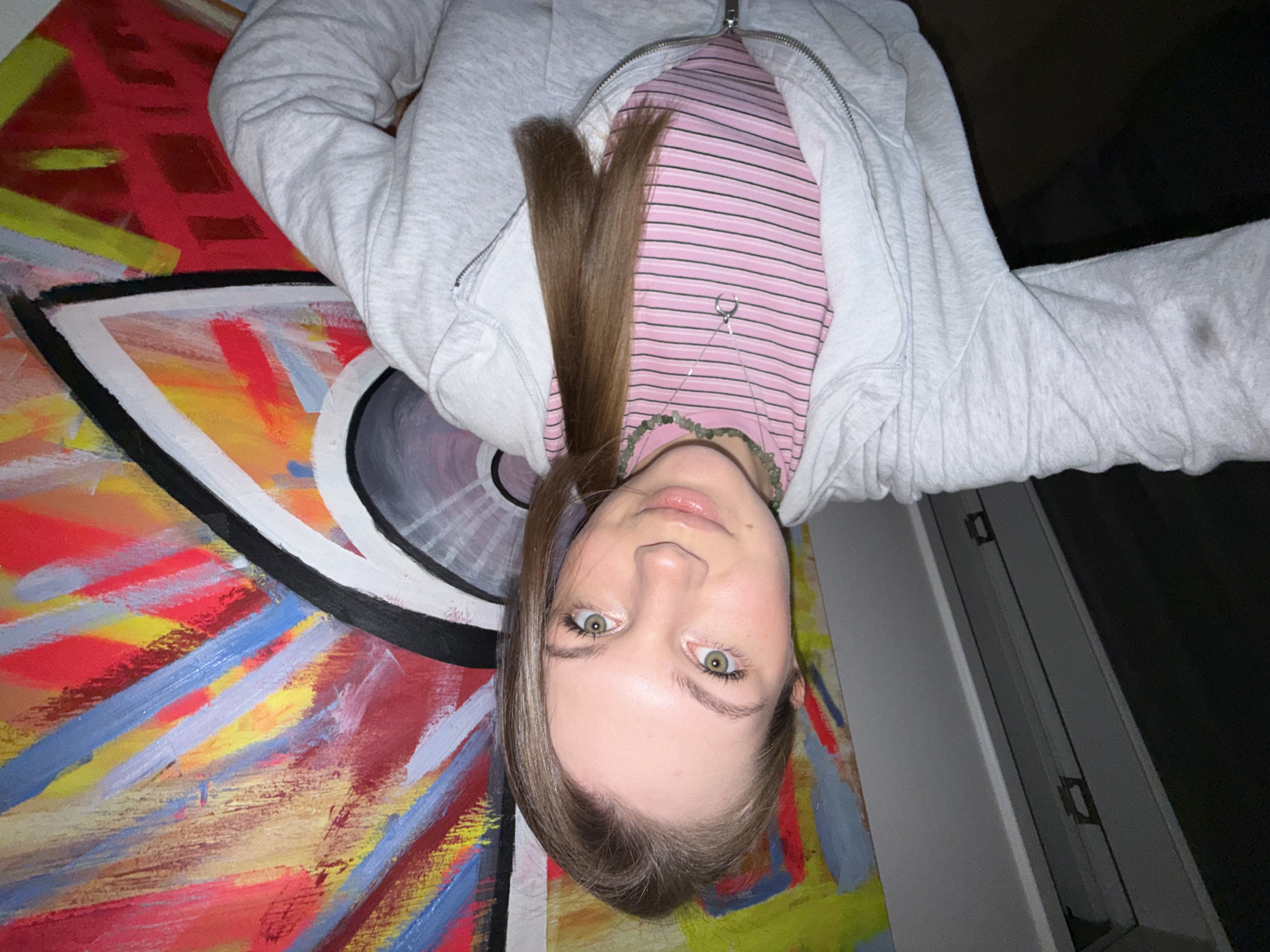 A person is lying down in front of a colorful abstract painting. The person has light skin, green eyes, and light brown hair. They are wearing a pink and white striped shirt, a gray zip-up hoodie, and a green beaded necklace. The painting is on a surface next to a white wall. The background includes a dark area, possibly a window covered with a dark drape. There is no evidence in the photograph to suggest this photograph was taken in Vozdovac Urban Municipality, Serbia.