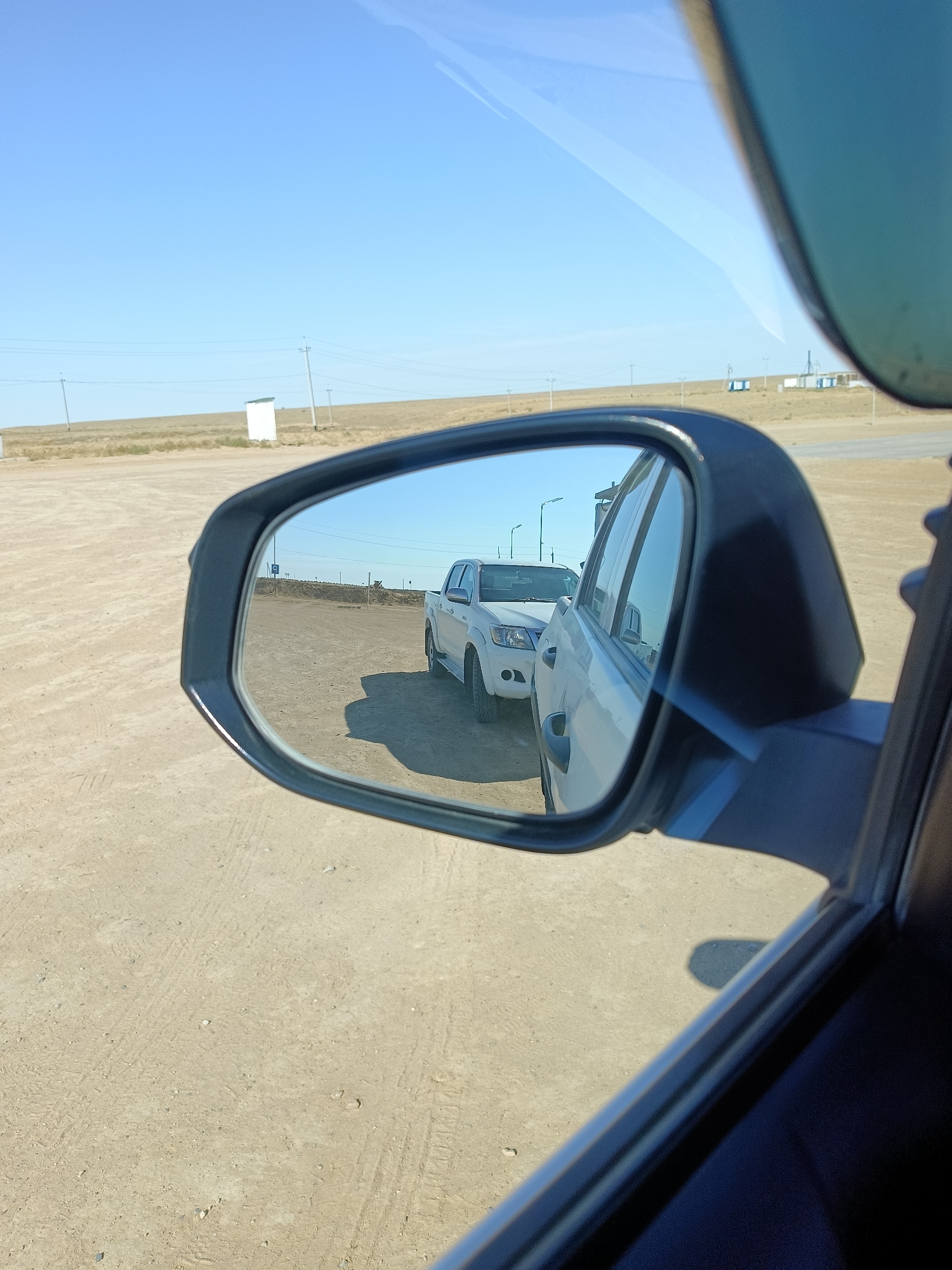 In this image, taken on a sunny day, a white pickup truck is seen in the reflection of a car's side-view mirror. The truck is parked on a dirt road with a pale, dry landscape surrounding it. In the distance, several utility poles with wires and some small white structures are visible against a clear blue sky. The scene suggests an outdoor location, possibly in a rural or undeveloped area. The image appears to be taken from inside a vehicle, looking outwards through the side window and into the side mirror. The environment is consistent with the arid or steppe regions of Kazakhstan.