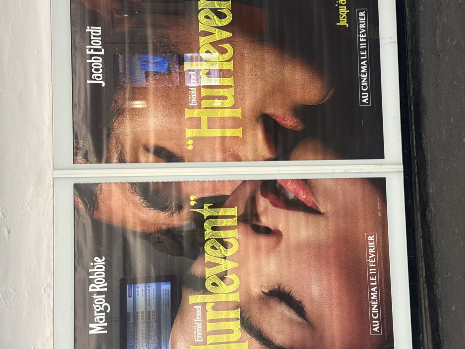 Two vertical, glass-fronted display cases, side-by-side, exhibit promotional movie posters. The posters collectively feature a close-up image of two human faces in intimate proximity. The face on the left is identified by superimposed white text as "Margot Robbie," depicted with closed eyes and a partially open, red-lipped mouth. The face on the right is identified by superimposed white text as "Jacob Elordi," depicted with a partially open mouth, seemingly in profile, facing the first face.
Both posters prominently display the film title "Hurlevent" in yellow, ornate lettering, with "Emerald Fennell" visible above it. Below the primary image on both posters, the French phrase "AU CINÉMA LE 11 FÉVRIER" (In cinemas February 11th) is printed in white.
The display cases are framed in white. Above them, a textured, off-white ceiling or upper wall surface shows minor imperfections. Below the displays, a dark, granular ground surface is visible. Reflections are apparent on the glass of the display cases: on the left, a digital screen displaying a grid-like pattern (likely a ticketing or information terminal) is reflected, along with the blurry image of a person holding a phone, presumably the photographer. On the right, reflections of blue and white light sources are present.
The presence of French language text and the style of the display indicate a public advertising environment in Paris, France, most probably within a metro station or a similar urban public transport corridor due to the reflections of digital screens and the utilitarian architectural elements.