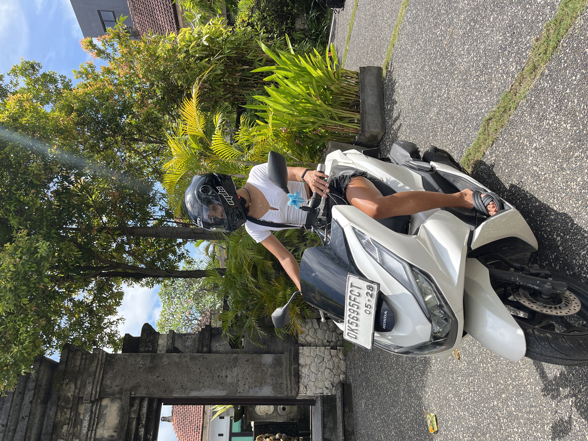 The image shows a person on a white Honda motorcycle. The license plate reads "DK5695FCT 05.28." The person is wearing a helmet, a white shirt, and dark shorts. The motorcycle is parked on a paved surface in a tropical outdoor environment with greenery and trees. The background features architectural elements that suggest the location is in Kuta Selatan, Indonesia.