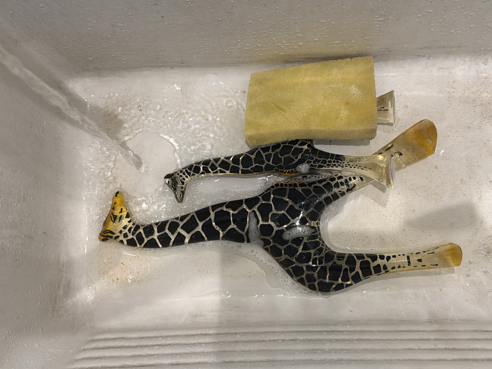 Two distinctly patterned objects, resembling stylized giraffes or possibly high-heeled shoes with a giraffe motif, are being washed in a white sink. One object is more prominently displayed in the foreground, showing a black and metallic gold or silver giraffe-like pattern across its body. Its front "legs" or toe area are a clear or amber-colored material, as are its rear "legs" or heel. A second, smaller object with a similar pattern is partially submerged behind it.
A yellow rectangular sponge rests near the top-right of the sink, also partially wet. Water is actively flowing into the sink from the left, creating ripples and bubbles, indicating a cleaning activity is underway. The sink's surface is white, with some visible water droplets on the upper walls and horizontal ridges on the bottom. The overall setting is an indoor utility space, likely a kitchen or laundry room, somewhere in Buenos Aires, Argentina. No people are visible, and there is no discernible text. The time of day and weather cannot be determined.