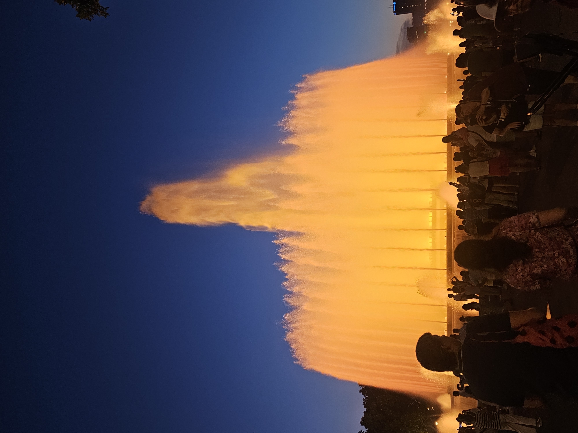 The image captures a vibrant performance by the Magic Fountain of Montjuïc in Barcelona, Spain. A large crowd of people is gathered, silhouetted against the dramatic display of water and light. The fountain jets water high into the deep blue twilight sky, illuminated with a warm, golden-orange glow. The water creates a luminous curtain, with intricate patterns and textures formed by the spray.
The scene suggests a public event or performance taking place in the evening. The sky transitions from a dark blue overhead to a lighter hue closer to the horizon, indicating dusk or early night. The presence of the crowd, facing the fountain, highlights the entertainment aspect of the scene. Some individuals are holding up what appear to be phones, suggesting they are capturing the spectacle. The overall atmosphere seems to be one of awe and enjoyment, with the colorful fountain serving as the central point of attention.
There are no easily decipherable text elements visible in the image. The focus is on the dynamic interaction between water, light, and the gathered audience. The fountain's performance is the main subject, with the crowd and the twilight sky forming the surrounding context.