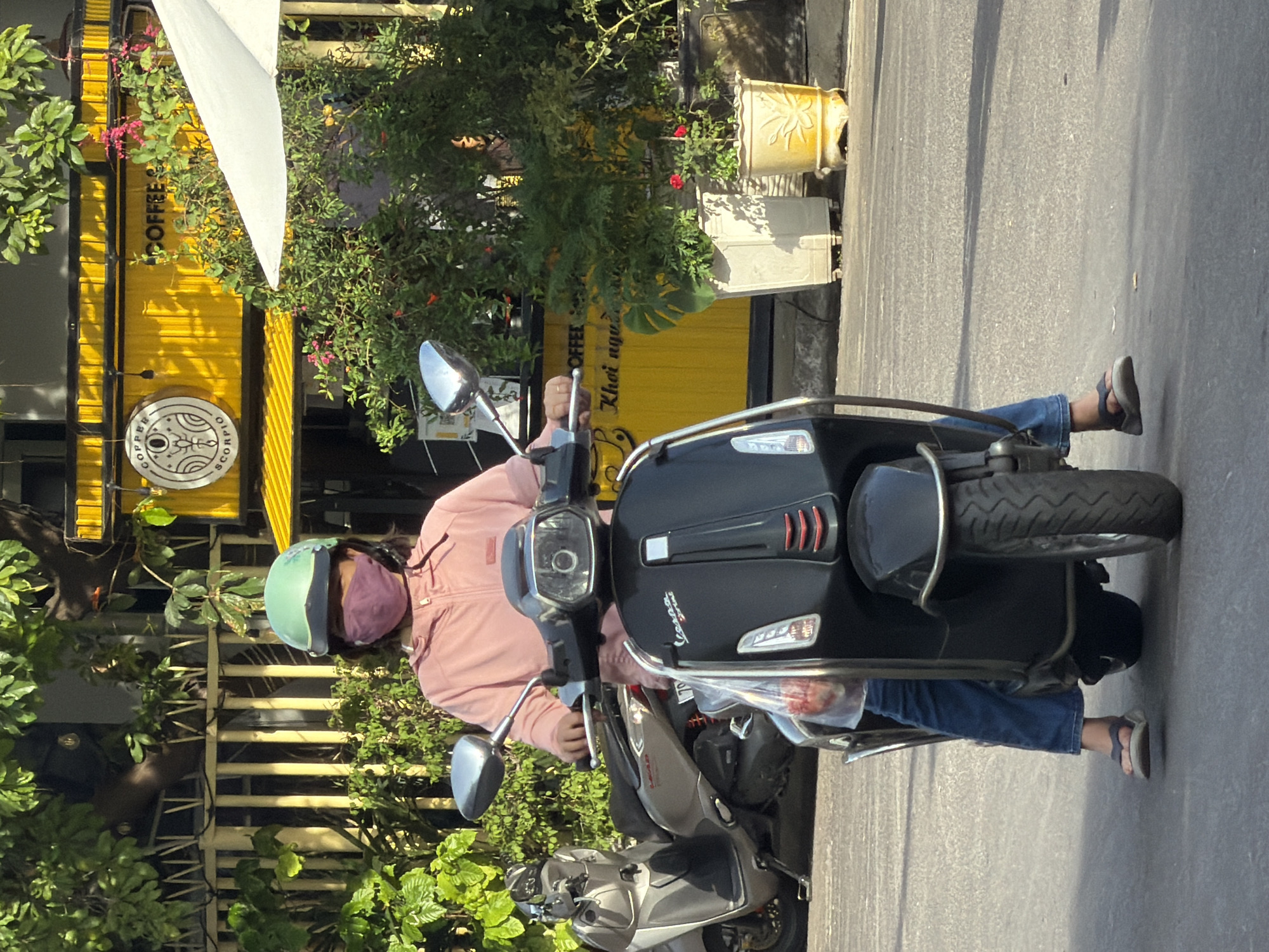 A person wearing a light pink hooded jacket, a purple mask, and a mint green helmet is sitting on a black Vespa scooter. The scooter has a clear plastic bag hanging from the handlebars. The person is wearing blue jeans and dark flip-flops. In the background, there is a yellow building with "COFFEE SCORPIO" signage and more text in Vietnamese. Lush green foliage and potted plants surround the building. The scene appears to be on a street in Nha Trang, Vietnam, indicated by the architecture, scooter culture, and signage.