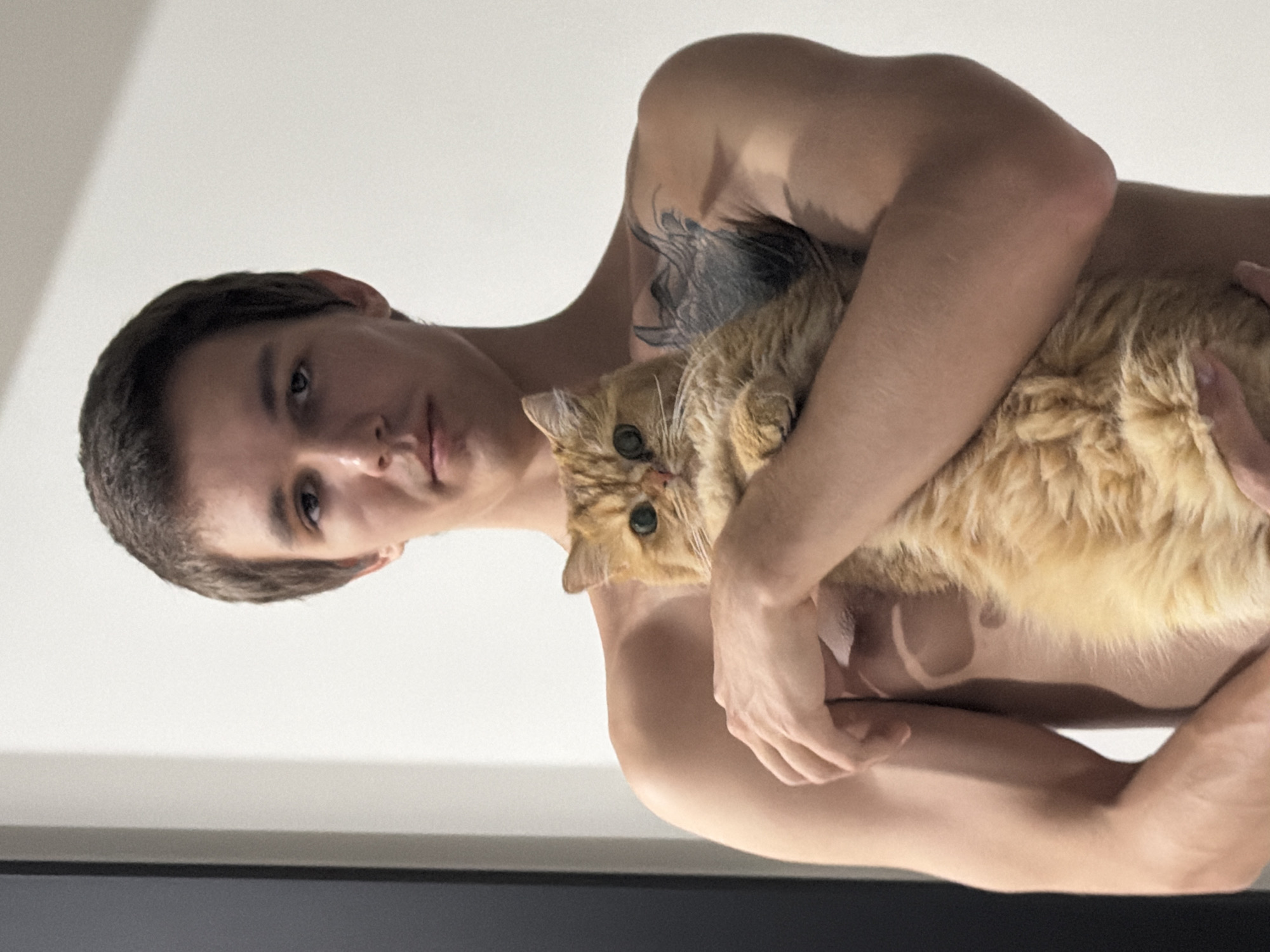 A young man with short brown hair and a tattoo on his chest is holding a fluffy orange cat. The man is shirtless and has tan lines visible on his chest. The cat is nestled in his arms, with its head resting on his arm. The background is a plain white wall with a dark vertical object on the left side, possibly a doorway or a piece of furniture. The lighting suggests an indoor setting. There is no discernible information about the location being Đà Nẵng, Vietnam.