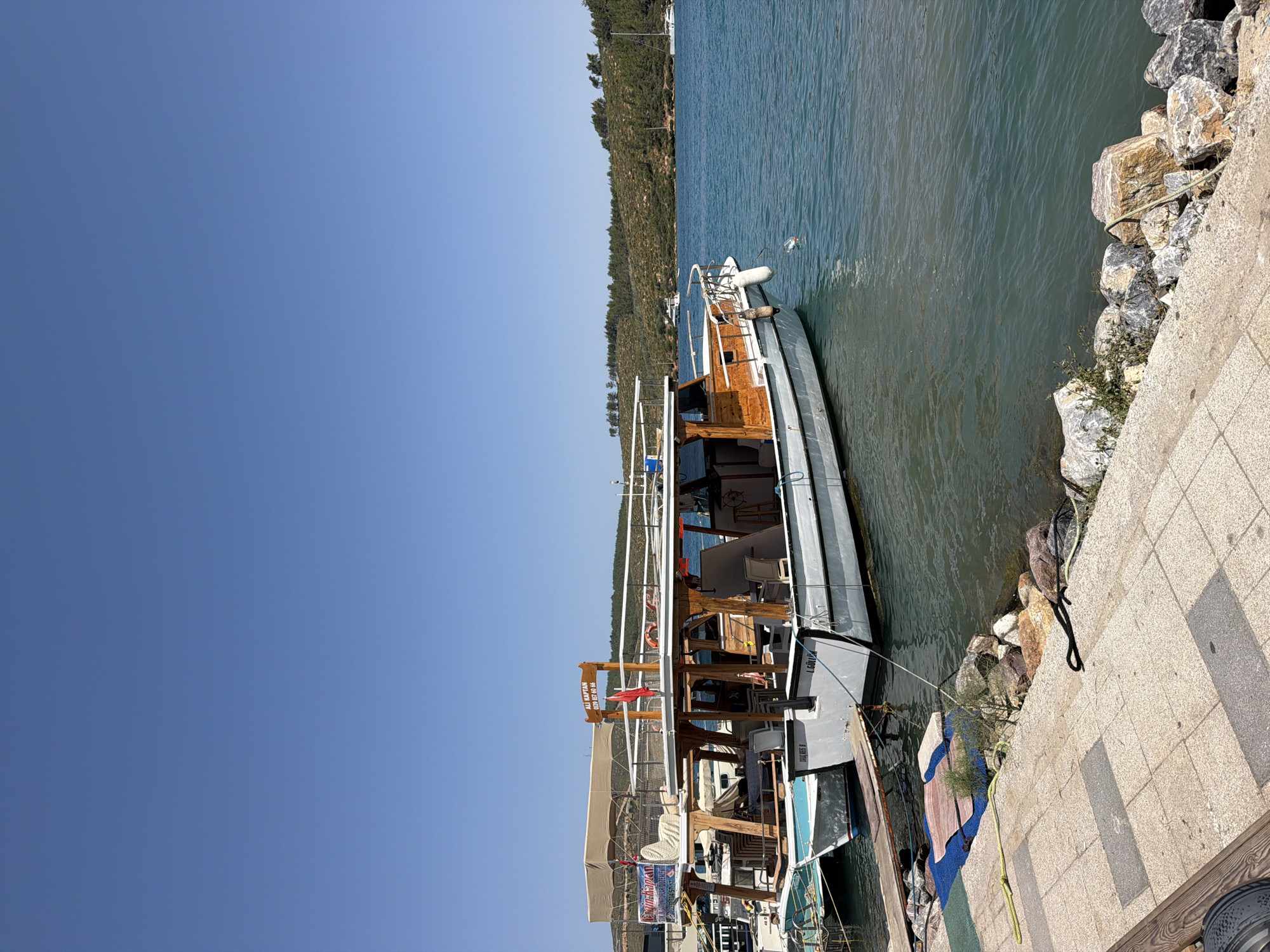 This is a wide, eye-level, outdoor, full shot of a dock in Bodrum, Turkey. In the foreground is a paved walkway next to a rocky shoreline. Beyond the shoreline are several boats docked in the clear, blue water. The largest boat, a white and wooden charter boat, is in the center of the frame. It has a covered upper deck with seating. The boat is named "ALI KAPTAN" with a phone number below it. The sky is clear and blue. The background shows a green, tree-covered hillside.