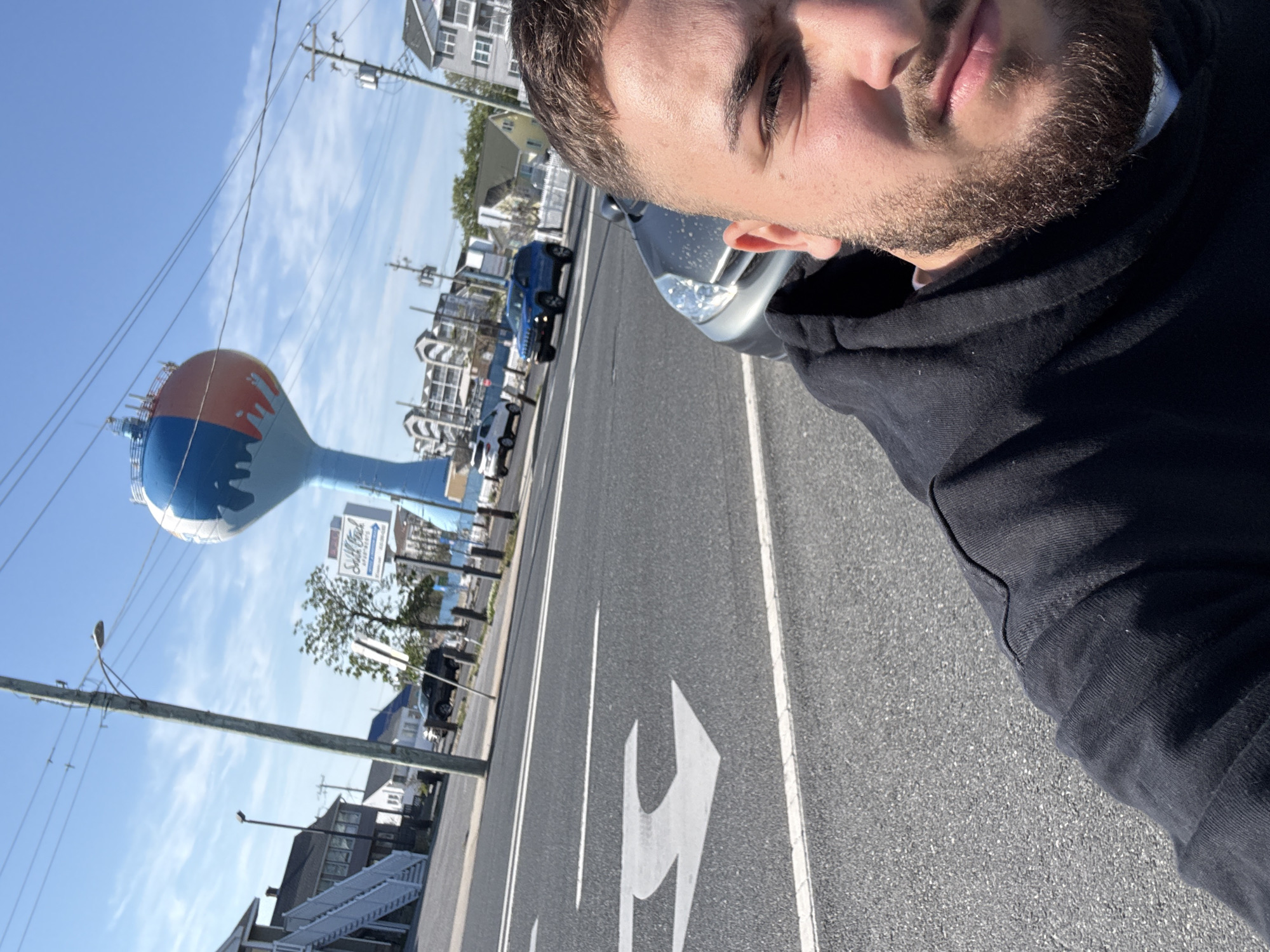 The image depicts a man in a black hoodie taking a selfie on a sunny day in Ocean City, New Jersey, United States. Behind him is a street with cars, buildings, and the iconic Ocean City water tower painted with an orange and blue design. The "Sift Shack" sign is also visible. The man is partially obscuring a silver car in the right corner of the frame. The road has directional arrows painted on it.