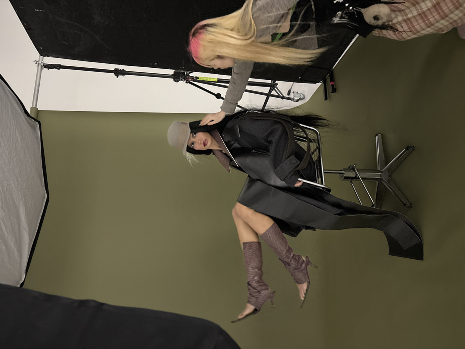 A woman sits backward on a rolling office chair, wearing a dark trench coat, gray hat, and brown high-heeled boots. Another person with blonde hair and pink roots touches the seated woman's head. The setup includes a large black screen above them, held by a metal stand. A large light diffuser is also visible in the scene, adding to the studio atmosphere. The background is a solid green wall.