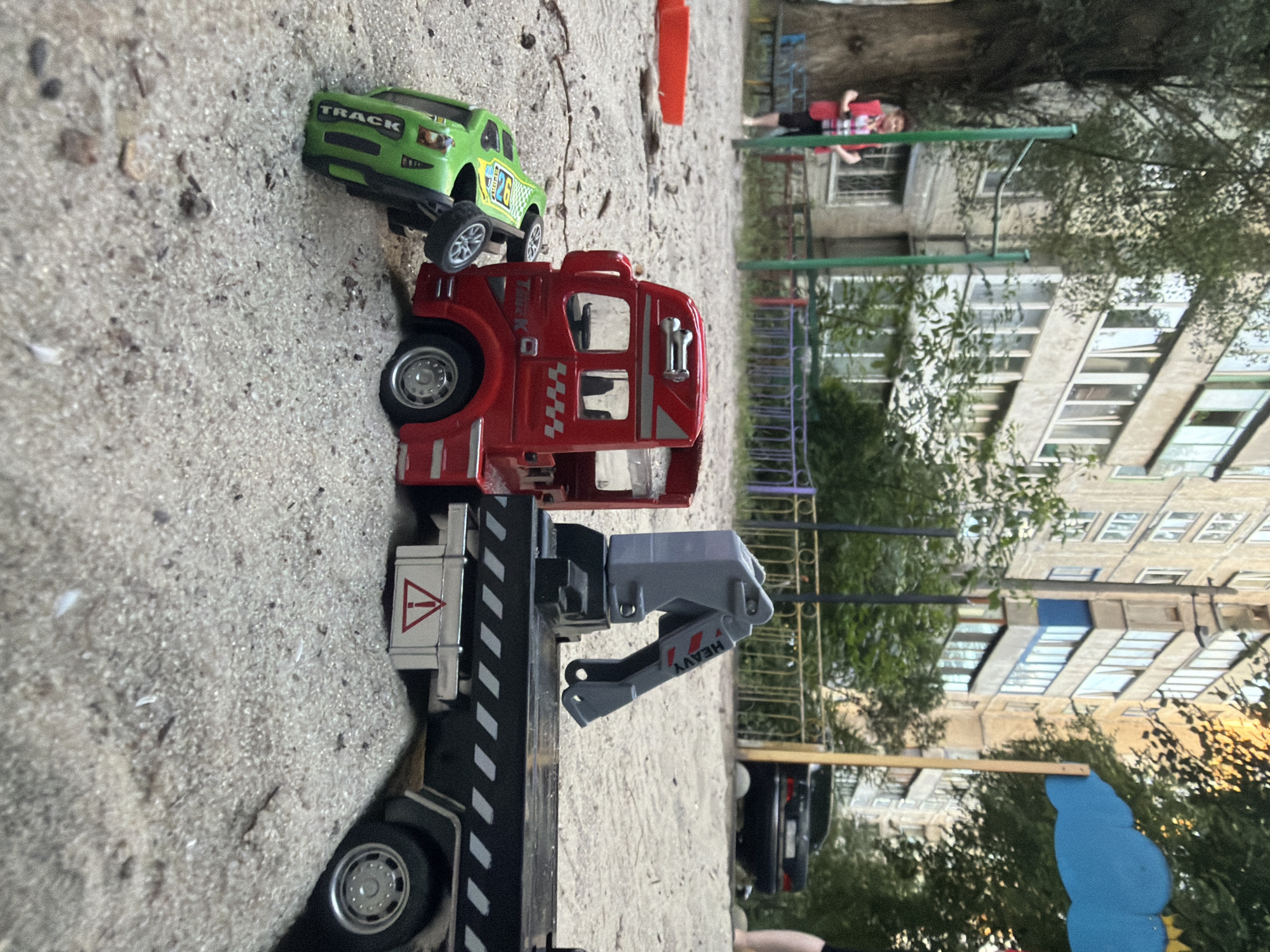 A low-angle medium shot captures a toy scene on a sandy playground, possibly in Kyiv, Ukraine. In the foreground, a red tow truck with a gray crane is parked on the sand. It is positioned behind a small, overturned green toy car. The car, labeled "TRACK," is on its side, with its wheels facing upwards. The tow truck has a reflective striping pattern on its black flatbed, and a red triangular warning symbol on a silver box below. The background shows a residential building with multiple windows and balconies, trees with green foliage, and a glimpse of a black car parked in the distance. A person in a red vest and plaid shirt is visible standing near a tree to the left, and another person's legs are visible on the far right. The overall scene suggests a playful, outdoor setting.