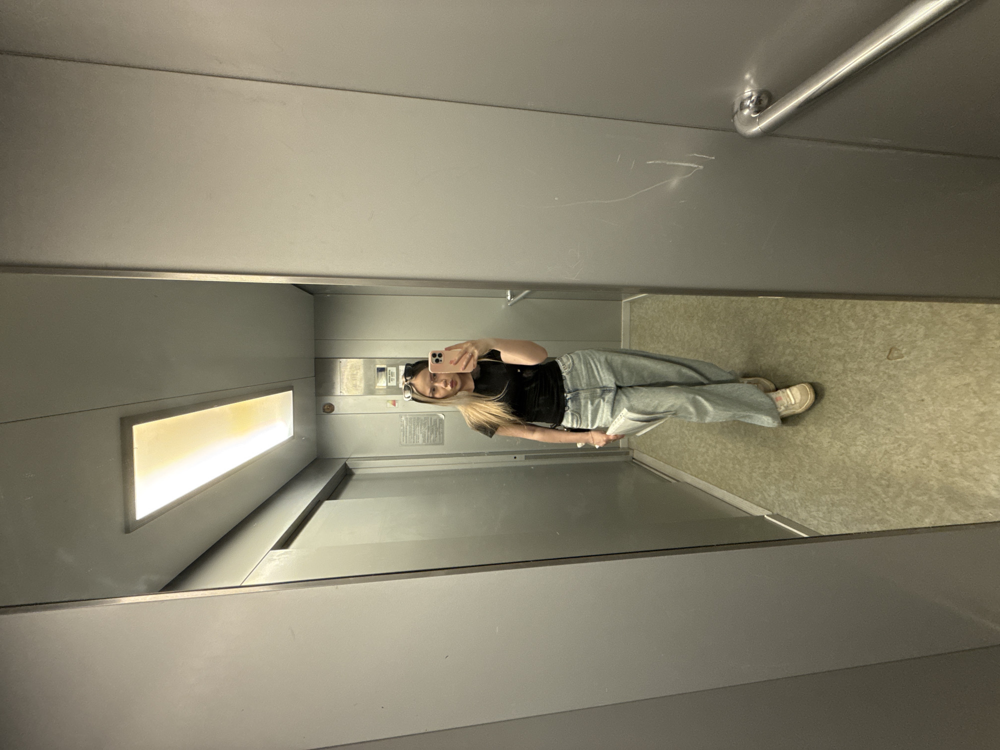 A photograph taken in an elevator shows a young woman reflected in a mirror. The interior of the elevator consists of grey metal walls. A window is present on one side of the elevator wall, with a light fixture inside. The woman is holding a smartphone in her hand, presumably to take the photograph. The floor of the elevator has a neutral color, speckled pattern. Based on the provided information, the location is presumed to be in the city of Chaikovsky, Russia.