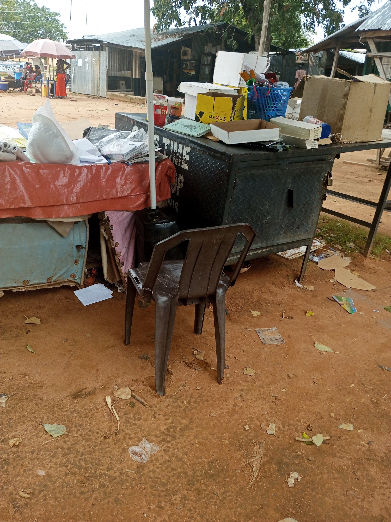 The image depicts an outdoor scene in Jalingo, Nigeria, likely a marketplace or roadside stall. The foreground is dominated by a brown plastic chair placed on reddish-brown dirt ground. Behind the chair is a stall with a metal counter on legs, covered with various items including boxes, papers, and a blue basket. Some of the boxes are labeled "NEXUS". A red cloth covers a section of the stall.
In the background, several corrugated iron structures suggest permanent or semi-permanent shops. People are visible in the distance, some under umbrellas, indicating a commercial area. The time of day appears to be daytime, with overcast or diffused lighting, suggesting it might be a cloudy day or late afternoon. The overall impression is that of a busy, informal trading environment. There are scattered leaves and some debris on the ground, typical of an outdoor setting in a tropical region. The structures and the presence of people suggest this is a community gathering place for commerce.