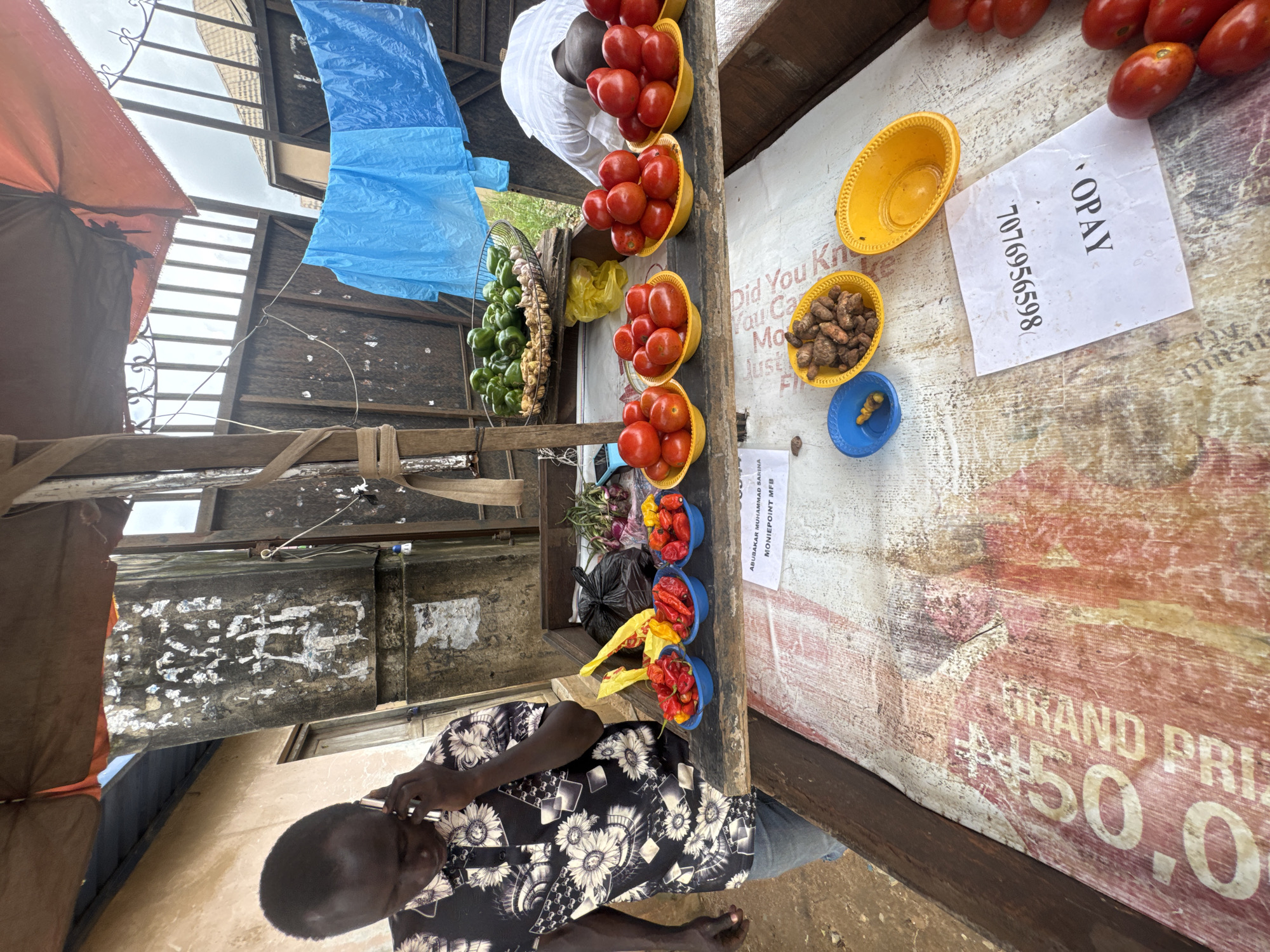 An outdoor market stall is set up in Ihiagwa, Nigeria. A dark-skinned man wearing a black and white floral shirt and blue jeans is on the phone, standing to the left of the stall. In front of him, on a wooden table, are several bowls of chili peppers. Behind the chili peppers are a black plastic bag, some onions, and a basket of green bell peppers and ginger. To the right of the chili peppers, on the same wooden table, are several bowls of tomatoes. Below this table is another wooden surface displaying a colorful, worn banner with large lettering and a paper slip with "OPAY" and a phone number printed on it. In the background, a blue tarp hangs and a metal fence with vertical bars is visible. The stall is covered by a rustic, orange and brown awning.