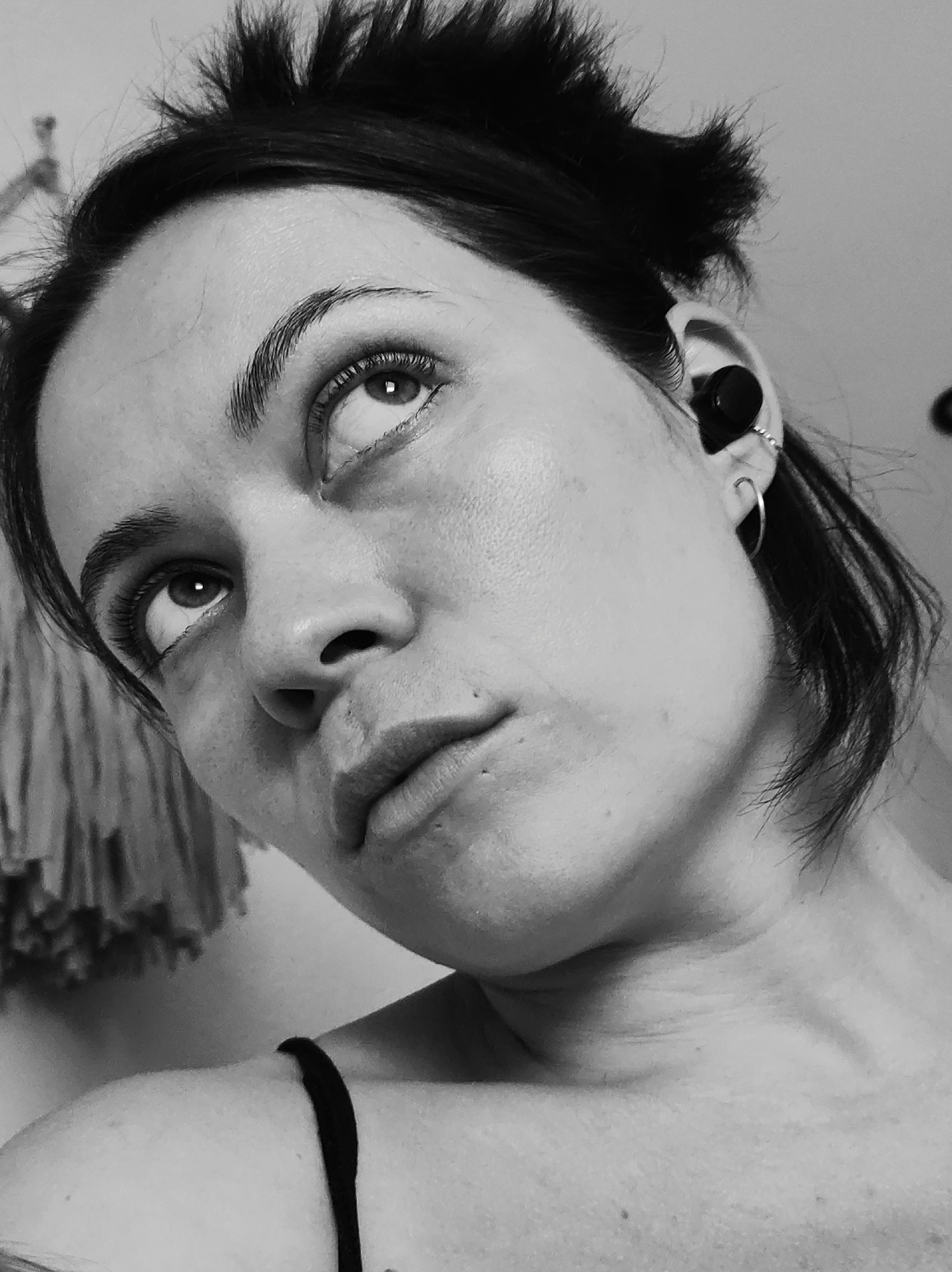 A black and white, low-angle portrait depicts a woman gazing upwards. She has dark hair, some of which is loosely styled upwards. She wears a small black earbud in her right ear, along with two hoop earrings. The woman's facial features are clearly visible, and the lighting emphasizes the texture of her skin. A thin strap of a garment is visible on her shoulder. In the background, partially visible on the left side, is a textured textile hanging on a wall. There is no contextual information to suggest the image was taken in the city of Chaikovsky, Russia.