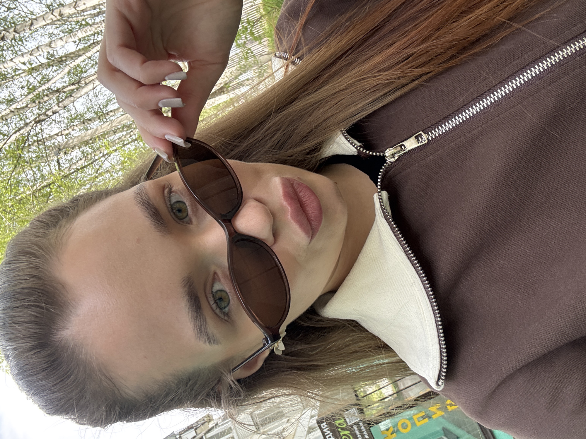 A fair-skinned woman is holding sunglasses and looking at the camera. She has light green eyes and light brown hair. She is wearing a brown jacket with a white collar. The background includes trees and a building. A sign in Russian can be seen in the lower right corner. Based on the writing on the sign, this is in the city of Chaikovsky, Russia.