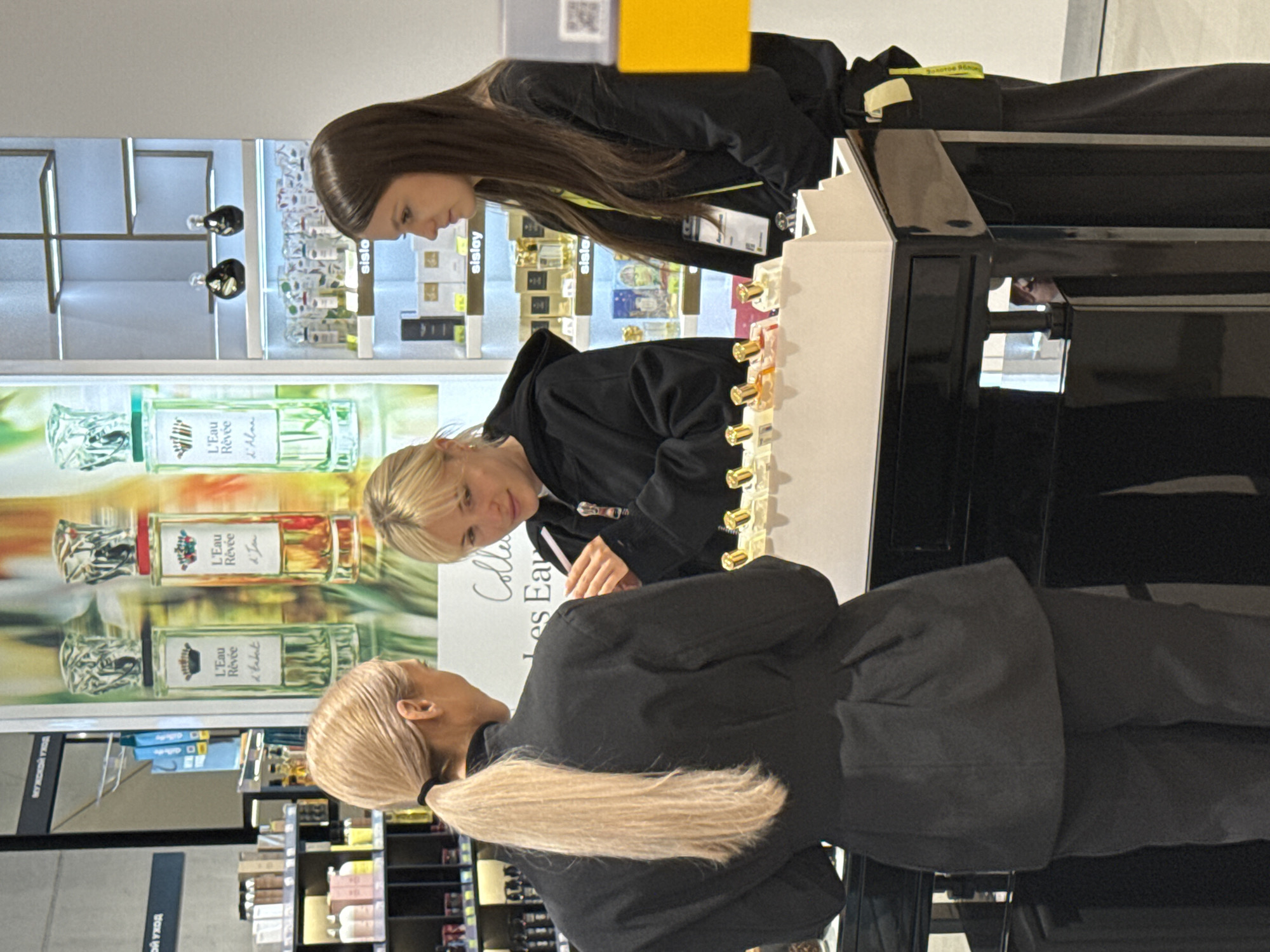 The image, oriented vertically, depicts three women engaged with a fragrance display in what appears to be a retail store, likely a cosmetics or perfume department in Kazan, Russia.
In the foreground, a woman with long blonde hair tied in a ponytail, dressed in a black jacket, stands with her back to the viewer. She is looking towards the display counter. Facing her, another woman with shorter blonde hair, wearing a black hooded top, leans over a sleek black and white counter, intently observing and possibly sampling small perfume bottles arranged in rows on its surface. Her hands are close to the bottles, indicating active engagement. To her right, a third woman with long dark hair, wearing a black top and a yellow lanyard, also looks towards the display, her gaze directed downwards at the products.
The background features large, brightly lit display panels showcasing prominent perfume bottles. Text visible on these displays includes product names such as "L'Eau Rêvée d'Alain" and "L'Eau Rêvée d'Tabat," along with what appears to be "Collection Les Eaux." The brand name "sisley" is clearly displayed on multiple smaller shelving units behind the women. Further in the background, shelves stocked with various products are visible, along with signs bearing Russian text, including "МУЖСКОЙ УХОД" (Men's Care) and "Золотое яблок" (Golden Apple), possibly indicating a store name or product category. The scene captures a typical retail shopping experience, with customers exploring fragrance offerings.