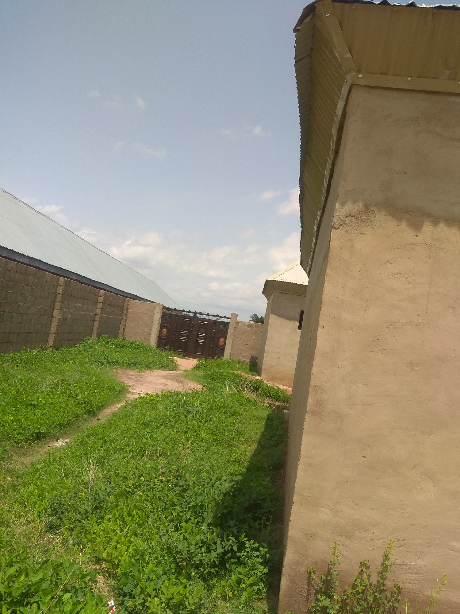The image depicts an outdoor scene in Jalingo, Nigeria, on a clear day with scattered clouds. The primary subjects are a wall and a gate, with a building in the background. The wall, constructed from concrete blocks, extends across the left side of the frame, topped with a corrugated metal roof. To the right of the wall is a large, ornate metal gate, dark brown in color with circular decorative elements. A path made of dirt or gravel leads towards the gate through an area covered in lush green grass and some small leafy plants.
In the foreground on the right, a building under construction or recently completed is visible. It has a rough, beige plastered exterior and a corrugated metal roof that angles upwards. Part of another building with a similar roof is visible behind it. The overall impression is of a secured property, possibly a residential or commercial compound, in a semi-rural or developing area. The lighting suggests it is daytime, likely late morning or afternoon, given the brightness of the sky and the shadows cast. There is no visible text in the image. The scene conveys a sense of quiet normalcy, with no active events taking place, focusing instead on the architecture and the surrounding environment.