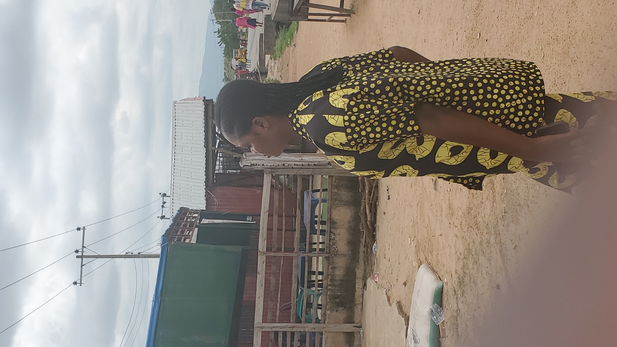The image shows a woman in a black and yellow patterned shirt standing in an outdoor environment. She is facing away from the camera, looking to her right. Behind her, there are wooden fences, tables, and chairs, suggesting an outdoor seating area or market stall. In the background, there are structures with corrugated metal roofs and power lines overhead. Further in the distance, there are more buildings, people, and a road with some vehicles, including a yellow tricycle. The ground is sandy and dusty, with some debris scattered around. The sky is cloudy. The overall scene appears to be a street or market area in a town or city. Given the provided context, this location is identified as the city of Jalingo, Nigeria.