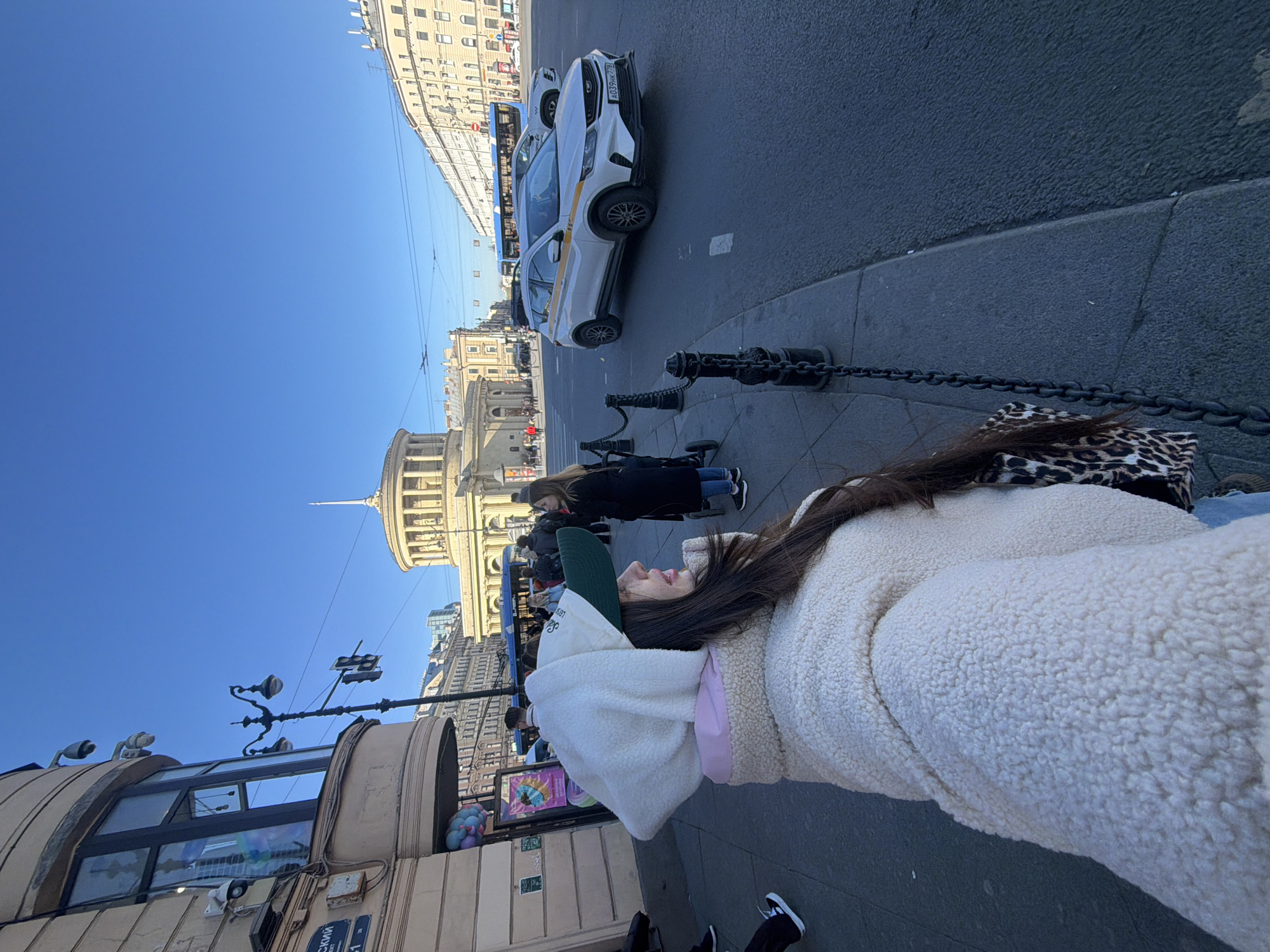A medium-shot photograph shows a person wearing a white, fluffy jacket and a white hoodie with a green baseball cap walking on a sidewalk in Saint Petersburg, Russia. The view is captured from a slightly low angle, looking up towards the sky and the surrounding cityscape.
The sidewalk is paved and features a black chain-link barrier. In the background, there is a street with vehicles, including a white taxi, and several people walking. Prominent architectural features include buildings with classical facades and a building with a dome. There is a streetlamp with a traffic light. The sky is clear and blue.