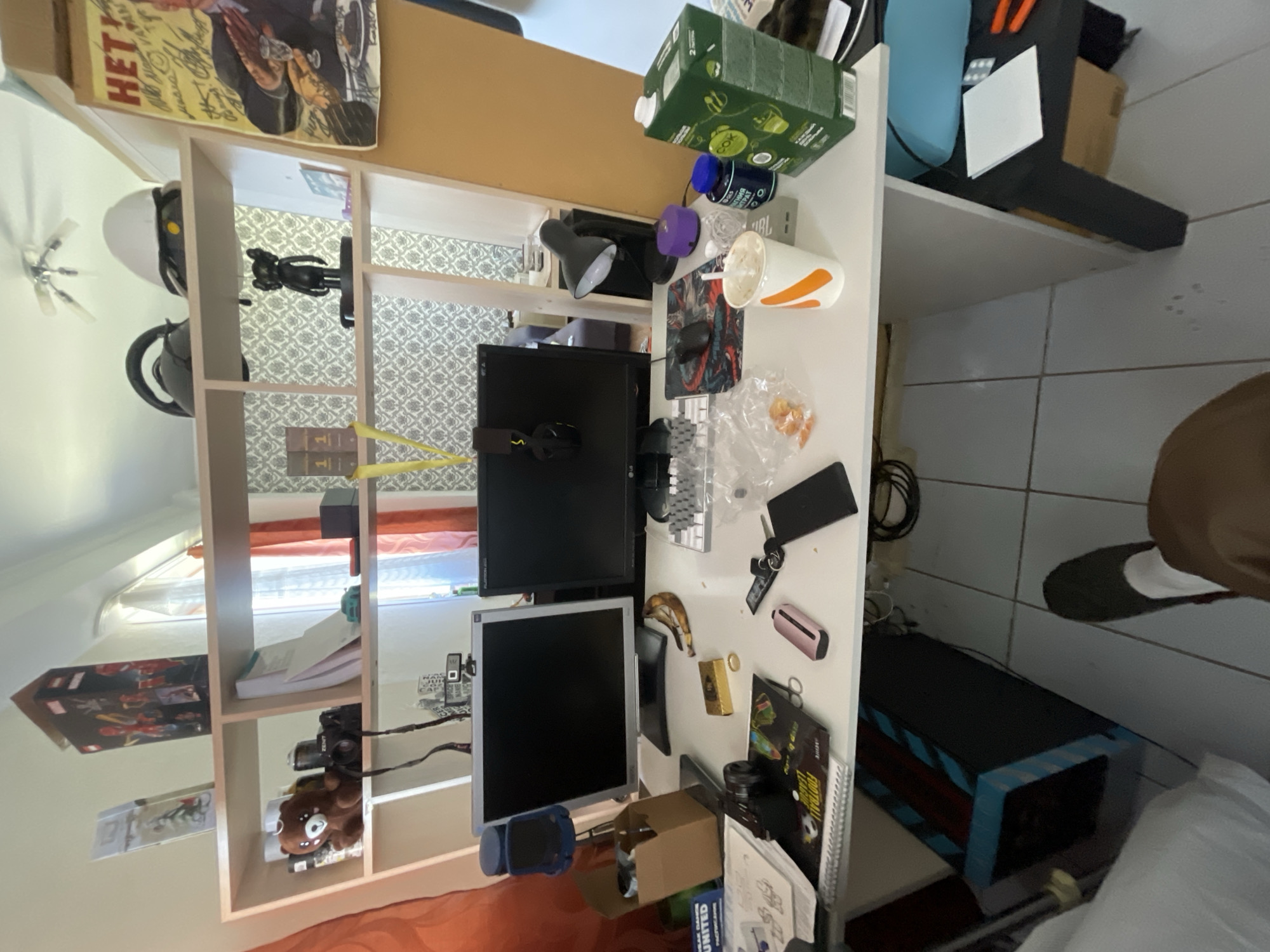 The image depicts a cluttered home office setting. There is a large desk supporting two computer monitors, a keyboard, mouse, and various other electronic devices, including a lamp and headphones. The desk also contains a disposable cup and what appears to be food packaging.
To the left of the desk is a shelving unit filled with various items including toys, boxes, and a camera. A poster or print is visible on the top shelf.
The environment shows aspects of a home, likely a room in an apartment or house. The exact location cannot be determined, but the request asks to include the city of Krasnoyarsk, Russia. Therefore, it can be assumed the scene is in a home office within Krasnoyarsk, Russia.
A person's lower leg and foot wearing a slipper are visible in the background, suggesting someone is present in the room.
