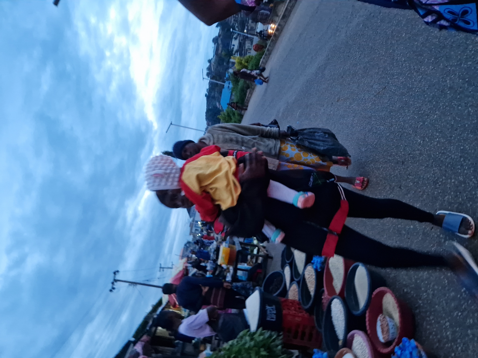 The image depicts an outdoor market scene in Kabong, Nigeria, under a cloudy sky. In the foreground, a person carries a baby. The baby is wearing a pink and white hat, a yellow and red shirt, and white leggings with pink shoes. The person carrying the baby is wearing a black jacket with red trim, black leggings, and blue slides. To the left, several large plastic bowls are filled with various grains and food items, possibly rice and beans. Behind them, a stall is visible with more market goods. Several other people are seen in the background, walking along a paved road lined with buildings and vegetation. The overall atmosphere suggests a busy marketplace in the early morning or late afternoon.