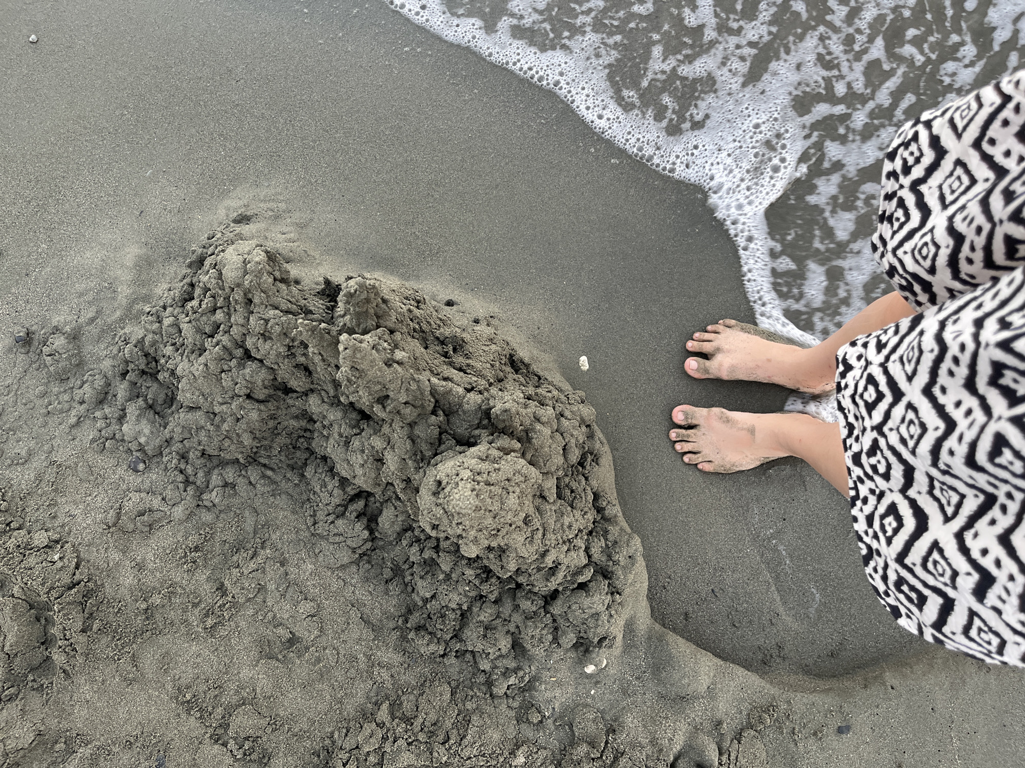 An overhead shot shows a person's feet standing on wet sand near the shoreline at the beach in Agios Tychona Community, Cyprus. The person is wearing a black and white patterned skirt. The sand is dark gray and wet from the incoming waves. There is a large pile of wet sand to the left of the person's feet. The water is shallow and foamy, with small waves lapping at the sand.