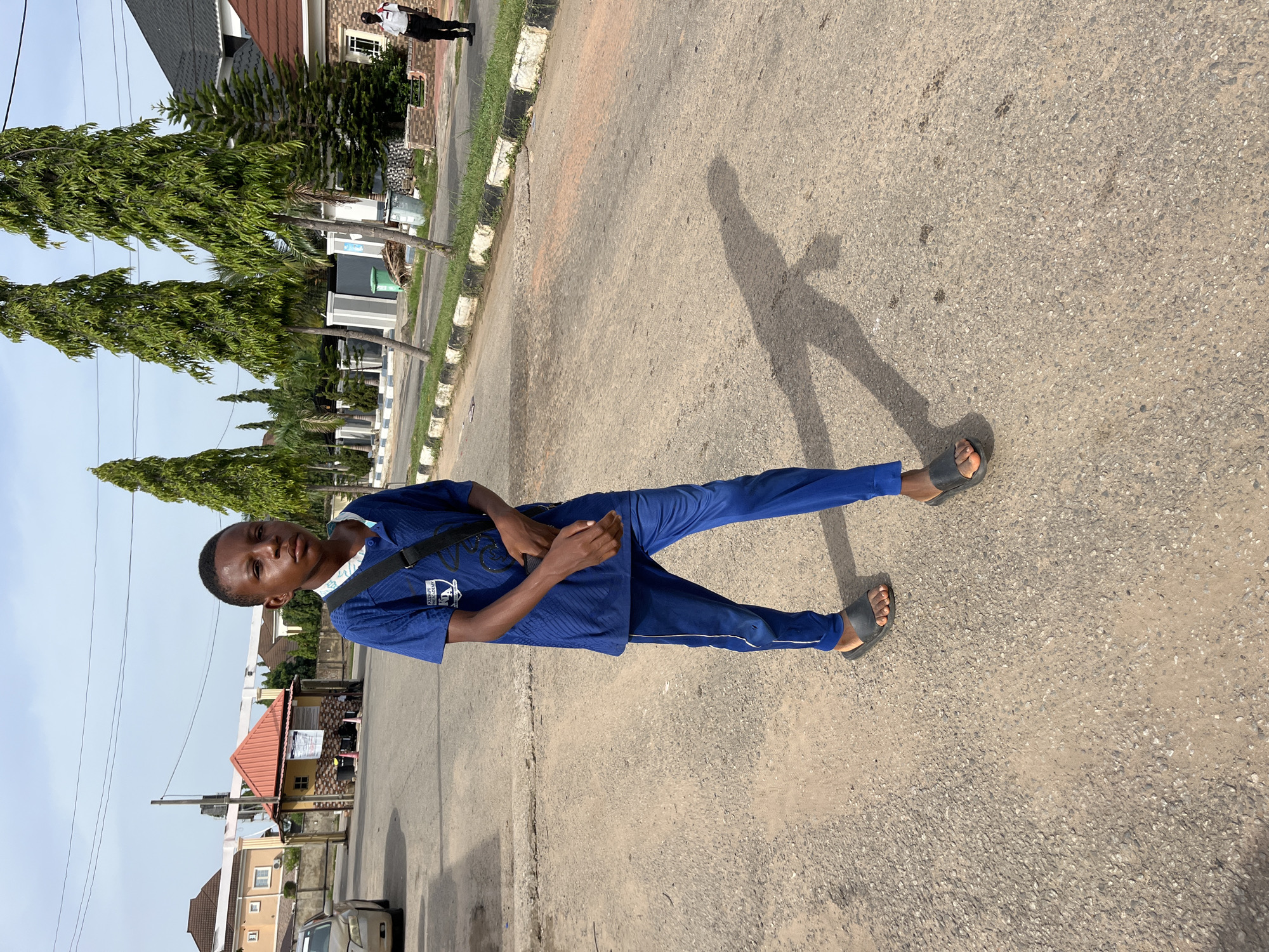 A young man stands on an asphalt road in Abuja, Nigeria. He wears a blue tracksuit and sandals, carrying a black shoulder bag. The background includes houses and trees. The weather is sunny, casting a long shadow.