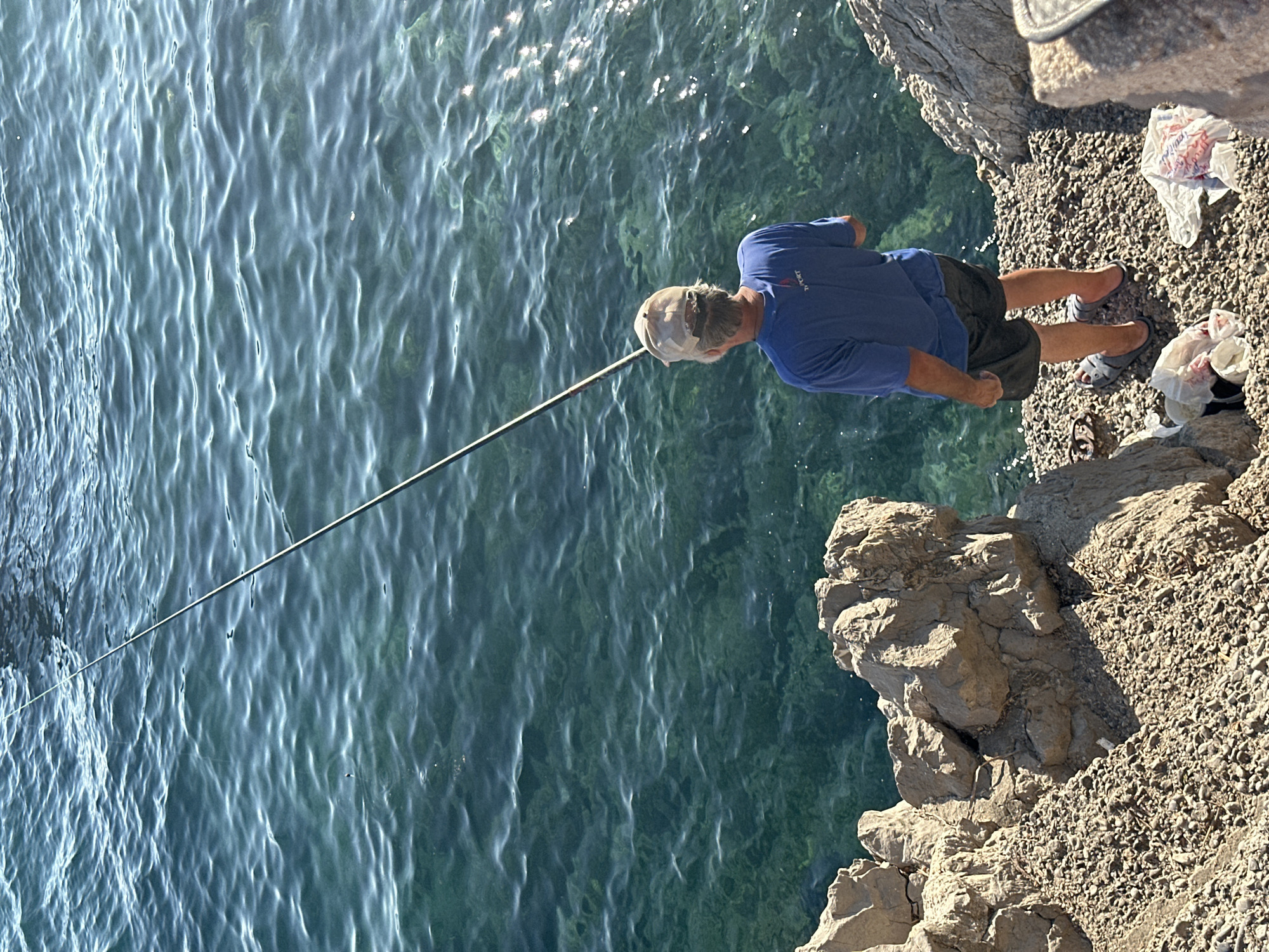 A man stands on a rocky shore, fishing in clear turquoise water. He is wearing a blue t-shirt, dark shorts, a beige baseball cap, and gray sandals. The fishing rod extends from his hands into the water. The shore is covered in gravel and large rocks. Several white plastic bags are scattered on the ground near his feet. The scene appears to be located in Budva, Montenegro, with the distinctive coastline and clear waters characteristic of the Adriatic Sea.