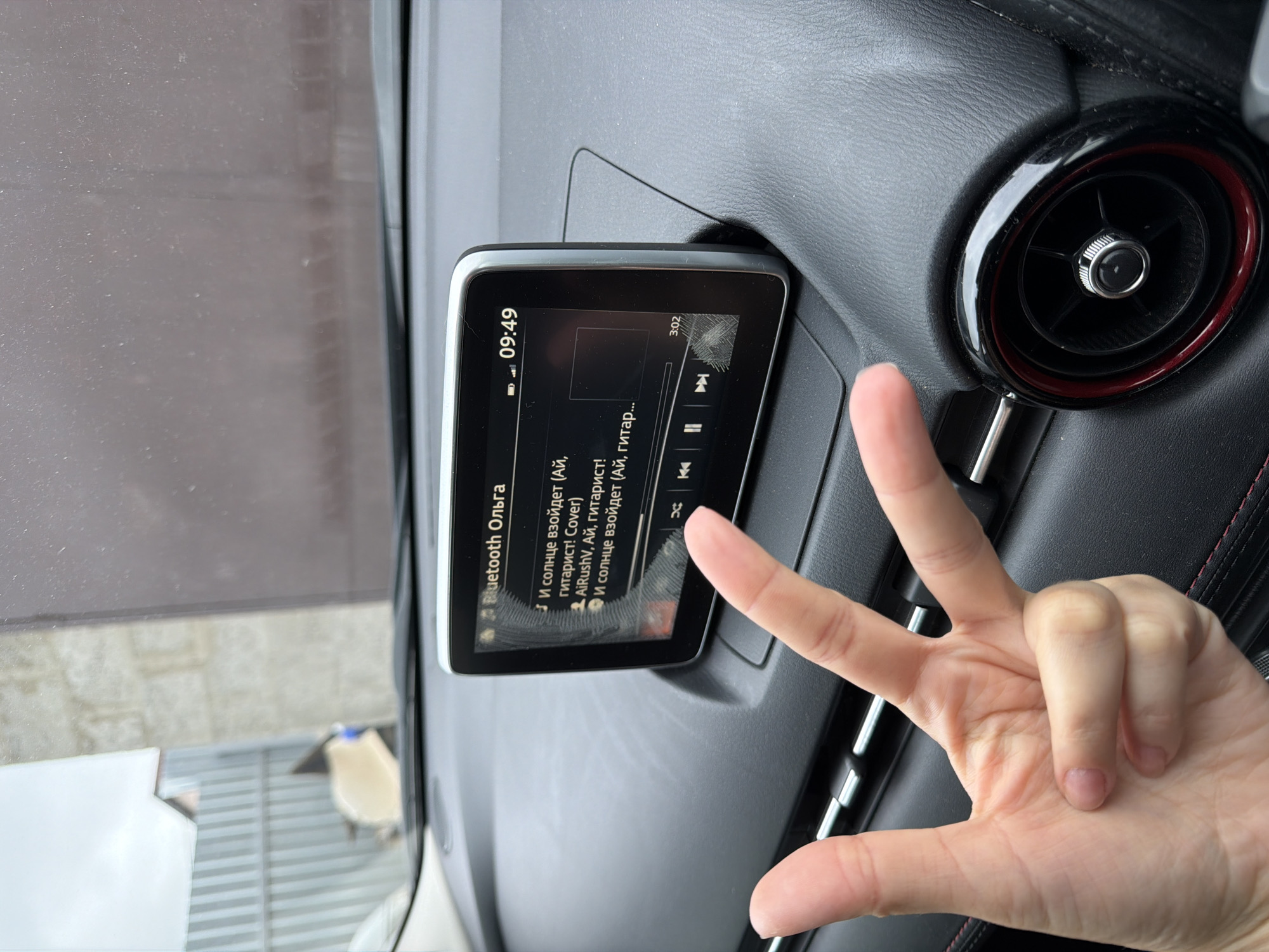 Here's a matter-of-fact description of the scene:
The image shows the dashboard of a car. A screen is mounted on the dashboard, displaying the name "Bluetooth Ольга" and a list of music tracks in Russian. A hand is making a "peace" or "victory" sign with fingers extended upwards in front of the screen. A circular air vent with a red ring is visible to the right of the screen. Through the windshield, a glimpse of an outdoor scene is visible, including a building and a sidewalk. The scene may be located in the city of Chaikovsky, Russia.