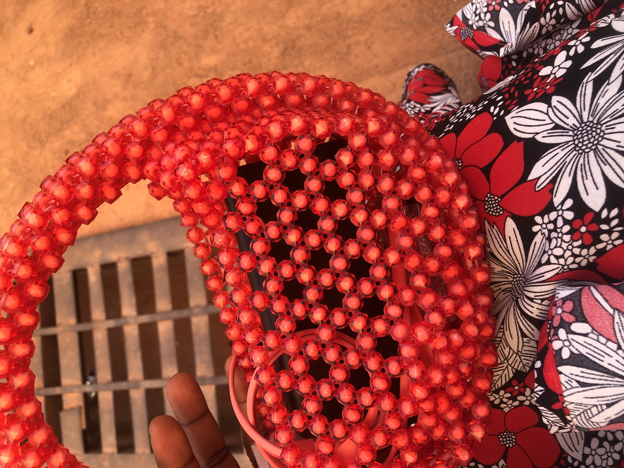 A close-up shot presents a red beaded bag, partially obscuring a black object underneath. The bag is constructed from numerous small, red beads joined together, creating a patterned, three-dimensional structure. A hand, possibly holding the bag, is visible on the left side of the frame. To the right of the bag, a floral-patterned fabric is present, featuring a mix of red and white flowers on a black background. The image suggests an outdoor setting with a grate visible in the background, while the red-brown surface behind the objects could be typical of an urban environment or building facade, possibly located in Rido, Nigeria.