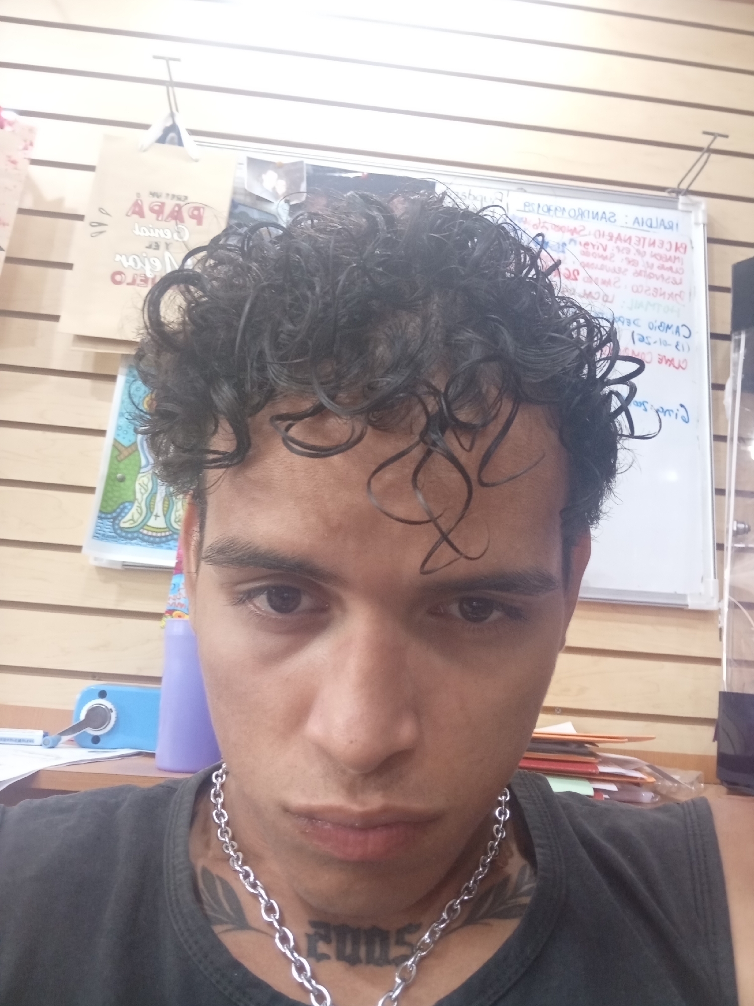 A close-up selfie shows a person with dark curly hair, brown eyes, and a silver chain necklace with a tattoo visible on their neck. The person is wearing a gray sleeveless shirt. The background includes a wall with horizontal wooden slats, a bag, a whiteboard with handwritten notes, and various objects on a table or desk. The location in the city of Juan Griego, Venezuela, is not evident from the image.