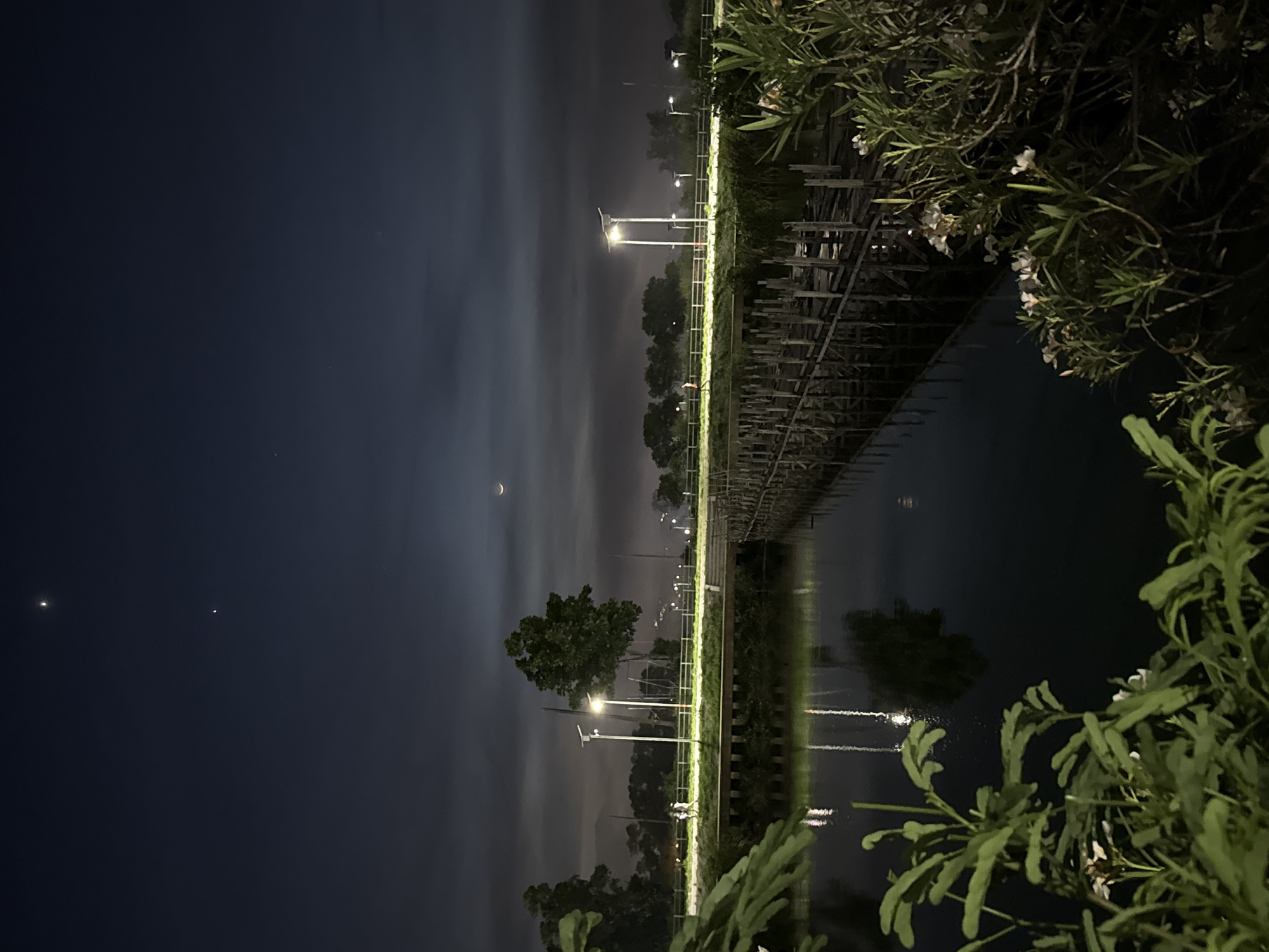The image depicts a nocturnal scene in Baan Chaweng, Thailand, likely a park or recreational area adjacent to a body of water. The sky is dark blue with some scattered clouds and a crescent moon visible. Two bright celestial bodies, possibly planets or bright stars, are prominent in the upper sky. A wooden walkway or bridge spans across the water, illuminated by intermittent lights along its path. The water reflects the lights and surrounding vegetation. On the bank, there are trees, streetlights with solar panels, and a low fence. Two figures are walking on a path beside the water, and another figure is visible further down the path. In the foreground, lush green foliage, including leafy plants and small white flowers, partially obscures the view of the water and bridge.