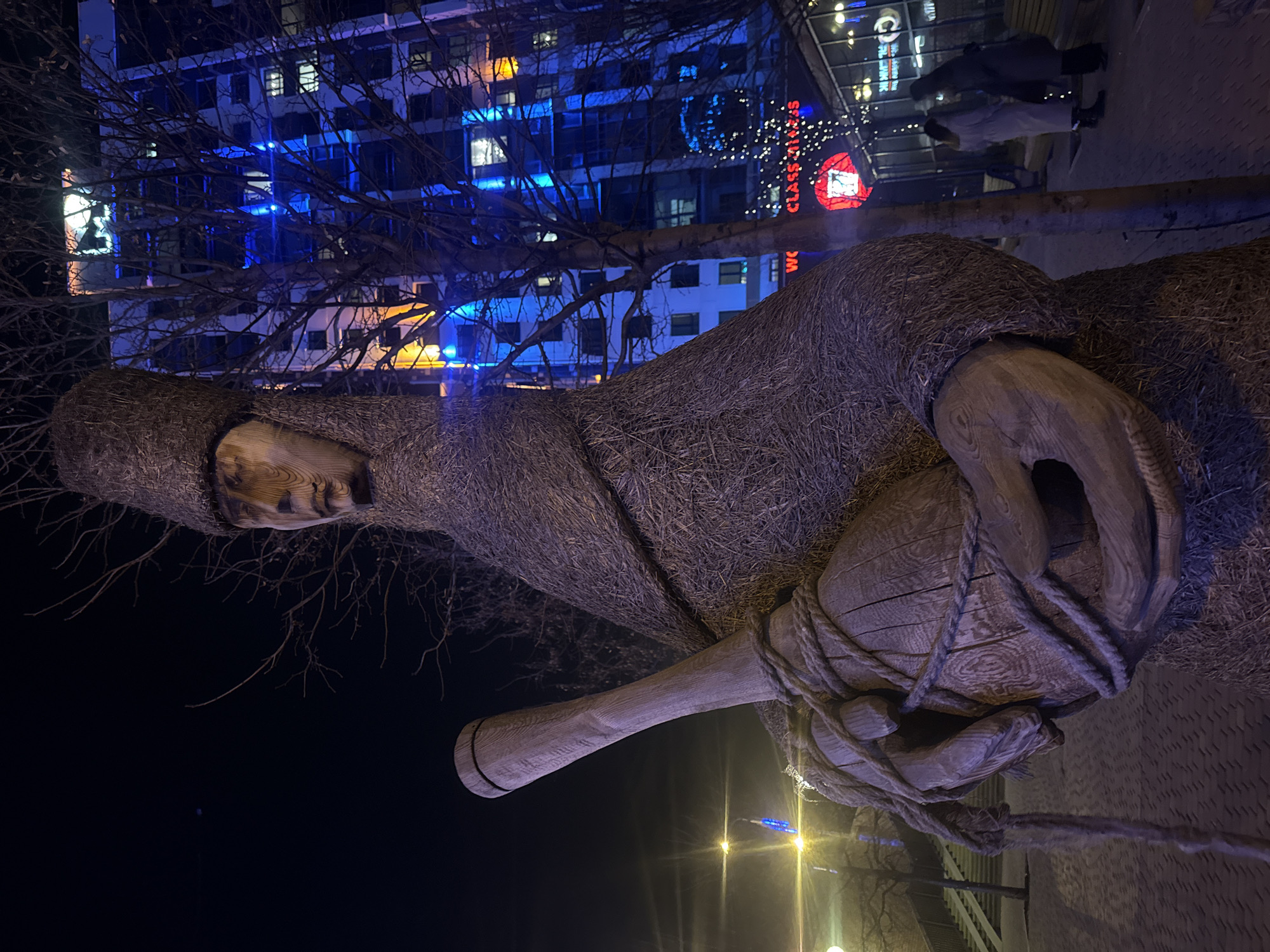 The image shows a nighttime street scene in Yekaterinburg, Russia. The primary subject is a large sculpture of a figure made of straw or a similar fibrous material. The figure is depicted holding a wooden object that resembles a bottle or flask, bound with rope. The background includes buildings with lit windows and decorative lights, suggesting an urban setting, potentially a commercial or public space. There are also silhouettes of people visible in the background, indicating pedestrian activity. The overall lighting is dim, with artificial light sources creating highlights and shadows.