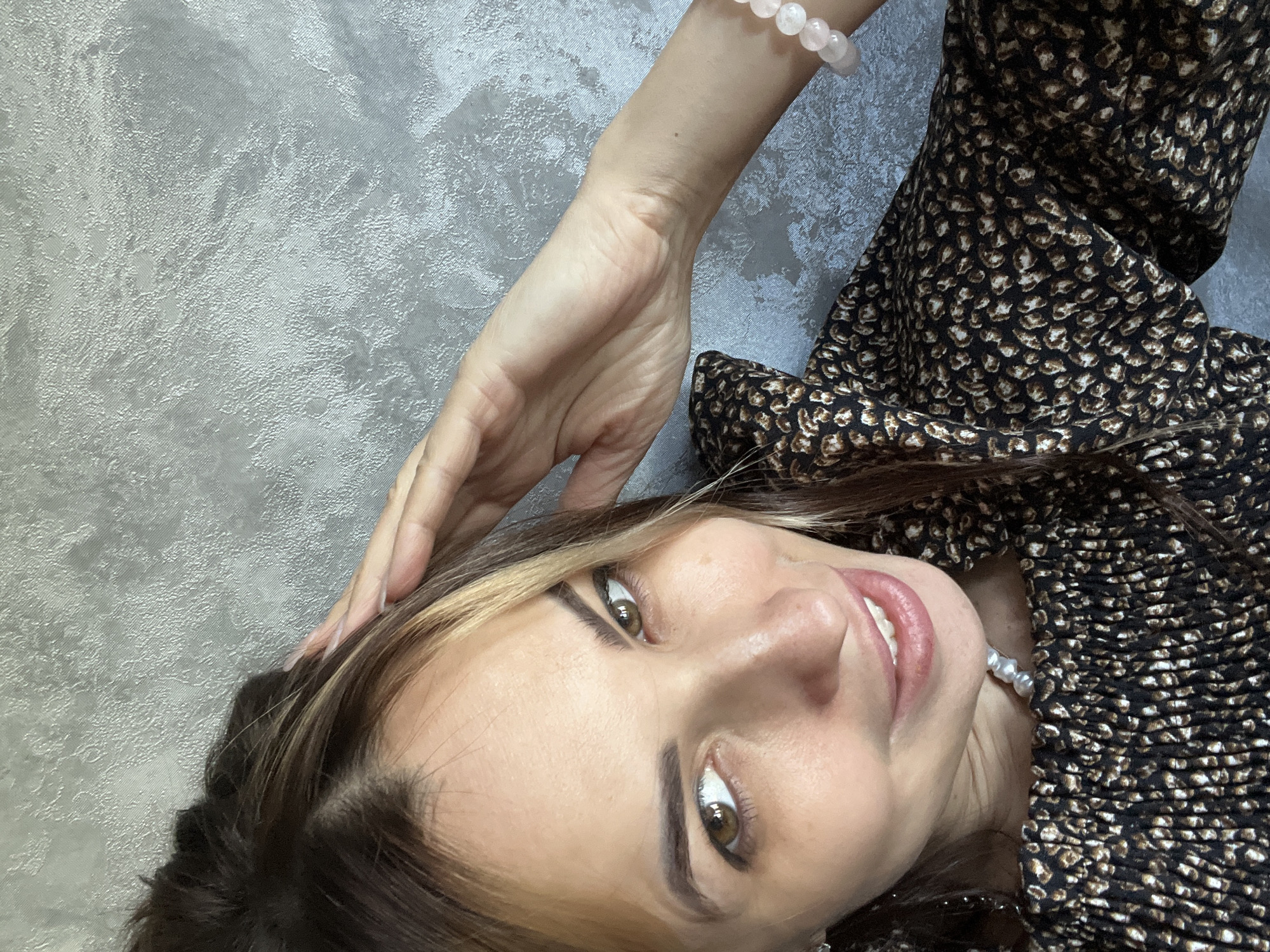 A woman with brown hair and hazel eyes is looking at the camera. She is wearing a black and brown patterned dress and a pearl necklace. She has a pink bracelet on her left wrist and her left hand is touching her hair. The background is a gray textured wall.