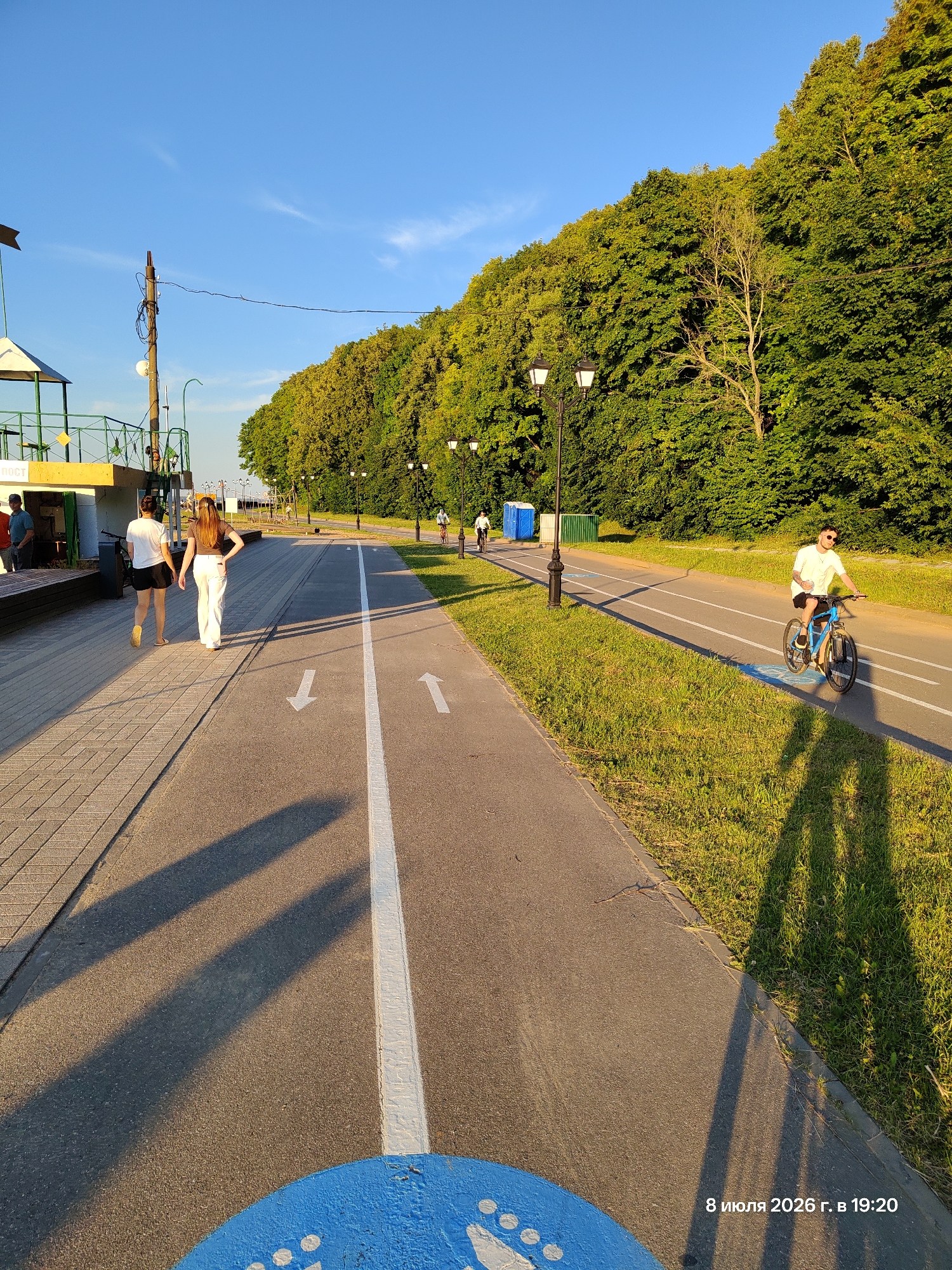 In an outdoor scene, two young women walk on a paved pedestrian path alongside a bicycle lane. The woman on the left wears a white t-shirt and black shorts, while the woman on the right is dressed in a brown top and white pants. They cast long shadows on the path, indicating late afternoon sun. A man on a blue bicycle rides in the opposite direction on the bike lane, also casting a long shadow. The path is bordered by a grassy area and a dense line of trees. A lamppost stands along the tree line. The sky is a clear, bright blue. The bottom right corner of the image features a timestamp: "8 июля 2026 г. в 19:20".
