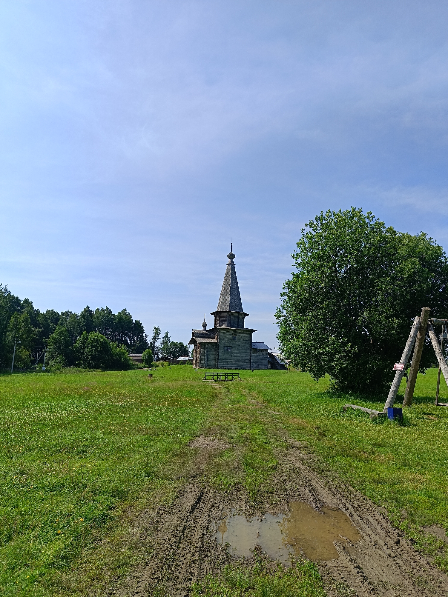 The image depicts a rural scene in Semenkovo, Russia, featuring a traditional wooden church as the central subject. The church, constructed of dark wood, has a distinctive tiered roof topped with a spire. In front of the church, a muddy track with tire marks and a puddle cuts through a vibrant green grassy field dotted with small yellow wildflowers. To the right of the church and field, a large, lush green tree dominates the landscape. Adjacent to the tree, a wooden swing set with two upright posts and a horizontal bar is visible. In the background, a dense line of green trees forms a horizon, with glimpses of some low-lying buildings peeking through. The sky above is a clear blue with sparse, wispy white clouds. The overall environment suggests a sunny day in a tranquil, natural setting.
