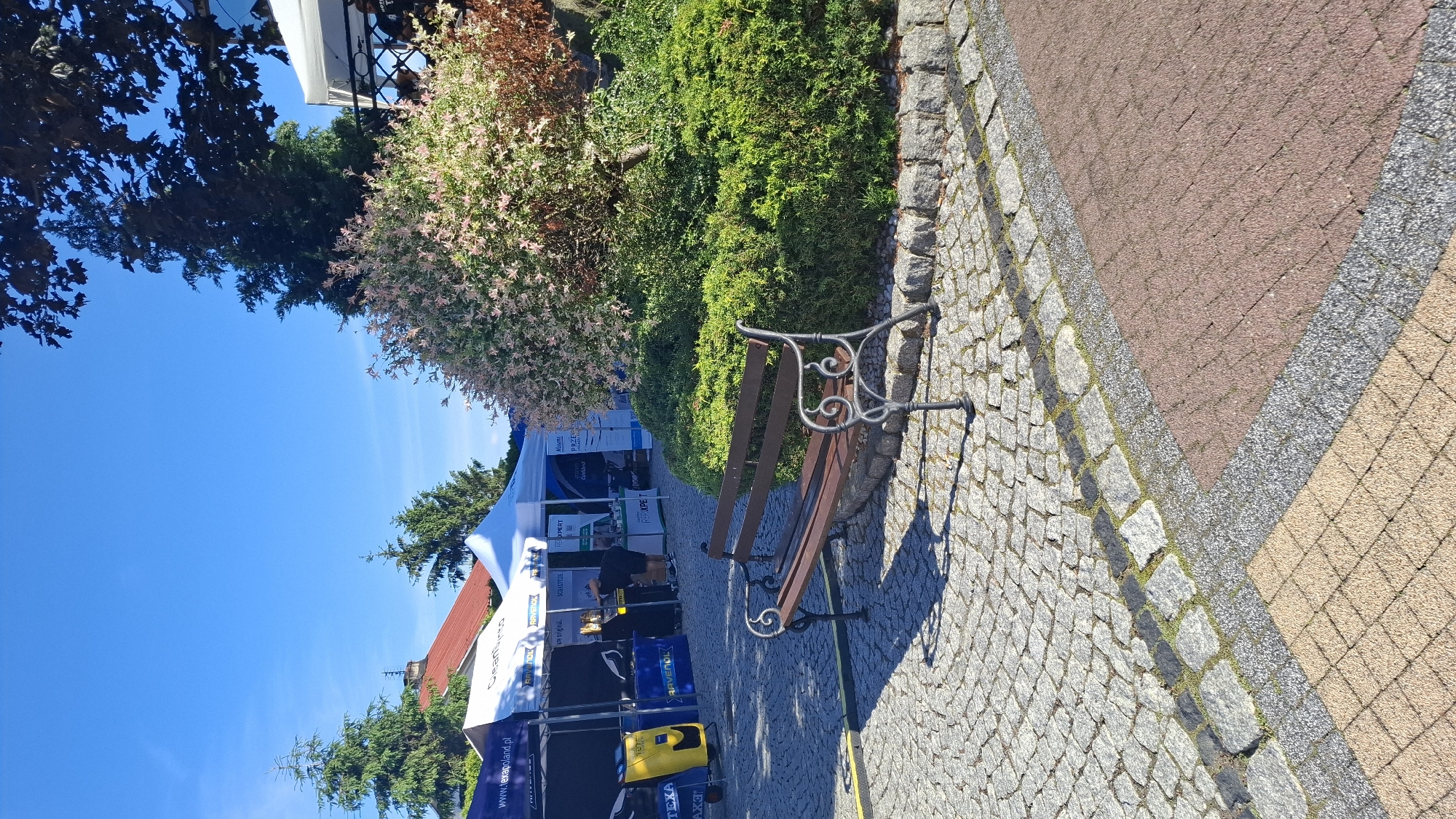 The image depicts an outdoor event or exhibition under a clear blue sky, likely on a sunny day. In the foreground, a stone-paved walkway leads towards several white tents. A park bench with a decorative metal frame and wooden slats sits on the right side of the walkway, casting a shadow. Behind the bench, there are lush green bushes and a tree with delicate pinkish-white blossoms.
The tents suggest a commercial or promotional event. Visible text on the tents and banners includes "RAVENOL," "Cleansint0," "www.texapoland.pl," "SCHAEFFLER," and "TEXA AX3." One tent displays what appears to be cleaning or automotive equipment, including a yellow device. A person can be seen inside one of the tents, possibly attending to visitors or setting up displays. The paving stones are varied, with cobblestones, red brick-like pavers, and grey gravel sections forming a distinct pattern.
The scene is set in a well-maintained outdoor area, possibly a park, town square, or a venue hosting an event. The overall atmosphere seems pleasant and engaging, with the bright sunlight and vibrant greenery contributing to a cheerful ambiance. The presence of promotional materials and visible text suggests a business or product launch, a trade show, or a local fair. The location context of Pokrzywnica, Poland, places the event in a specific geographical setting.