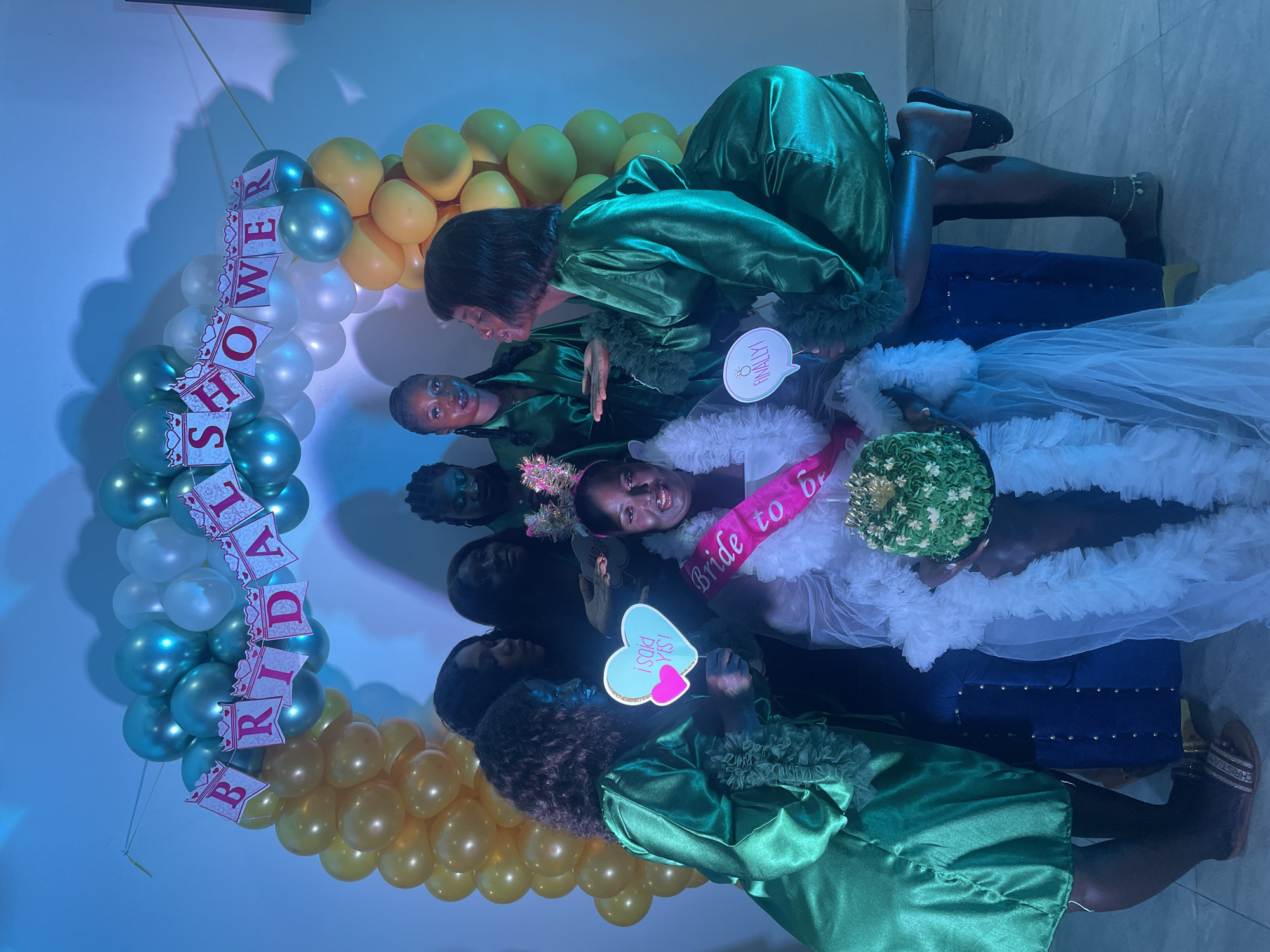 A group of women are gathered in front of a balloon arch and a banner that reads "BRIDAL SHOWER." The bride, wearing a white ruffled dress and a "Bride to Be" sash, is seated and holding a green cake. Other women, dressed in matching green satin robes, surround her, holding signs with phrases like "I SAID YES!" and "FINALLY!". The scene appears to be a bridal shower celebration. The environment is indoors, with blue lighting and a tiled floor.