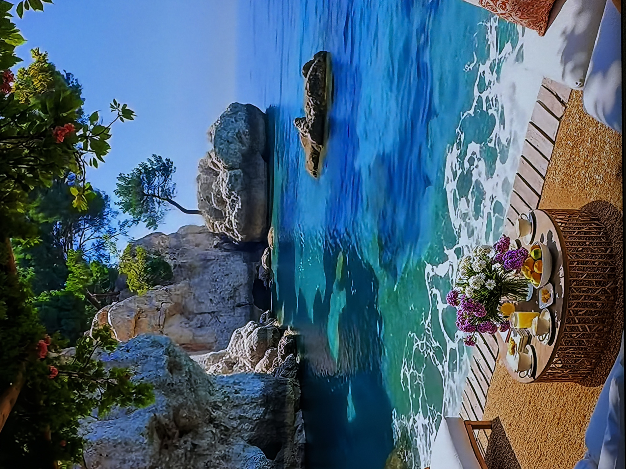 The image displays a vibrant, sunny coastal scene, likely a serene luxury resort setting. The perspective is from an elevated outdoor area, looking out over clear blue-green waters.
In the foreground, a round, woven table with a wooden surface is meticulously arranged for a meal. It features a lush bouquet of purple and white flowers in a vase, bowls of fresh oranges or mandarins, glasses filled with orange juice, and elegant teacups or coffee cups with saucers, alongside small plates that might hold pastries or snacks. Adjacent to the table, part of a wooden deck or platform is visible, leading towards the ocean, with soft white cushions and a textured brown rug suggesting a comfortable lounging area.
The vast midground is dominated by the sparkling, clear ocean, with gentle white foamy waves breaking along the shore and against scattered rock formations. Large, rugged boulders and rocky cliffs define the coastline, some partially submerged in the transparent water, revealing hints of the seabed.
On the left side of the frame, dense green foliage, including trees and bushes adorned with hints of pink or red blossoms, frames the view, adding to the lush natural beauty. A solitary tree is visible growing from the rocky terrain further in the distance. The sky above is a clear, brilliant blue, indicating bright, sunny weather.
No people are present in the image, emphasizing a sense of peace and an undisturbed, idyllic environment. The time of day appears to be morning or early afternoon, suggested by the bright light and the breakfast/brunch setup. There is no visible text in the image. The scene evokes feelings of relaxation, natural beauty, and a luxurious getaway.