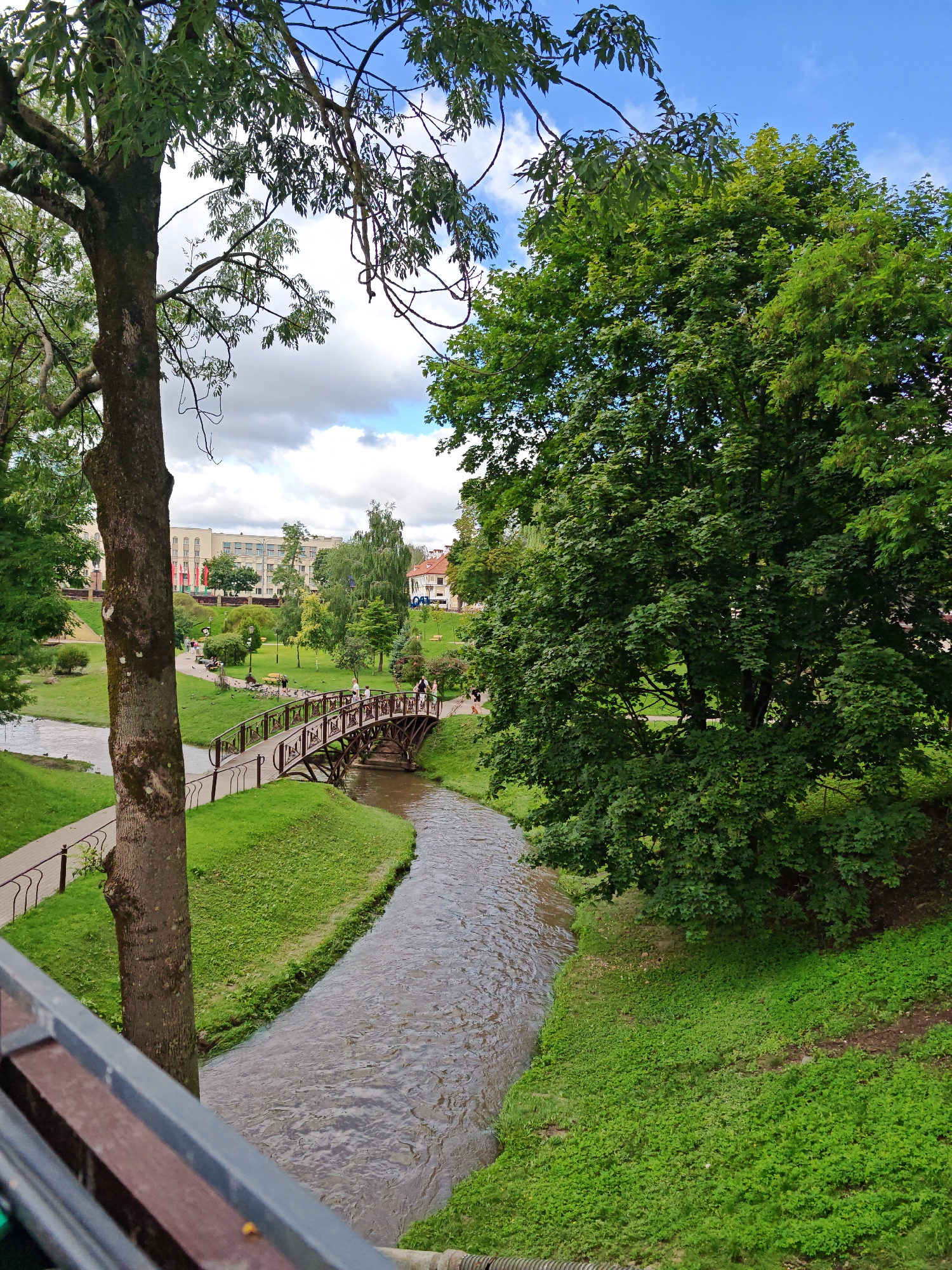 The image captures a scenic view of a park in Hrodna, Belarus, featuring a winding stream flowing through lush green grass. A charming wooden footbridge arches over the stream, with several people casually strolling across it. The park is adorned with mature trees, their verdant foliage providing a vibrant backdrop. In the distance, buildings with light-colored facades are visible, suggesting an urban setting adjacent to the park. The sky above is a mix of blue and white clouds, indicating fair weather. The foreground reveals a portion of a wooden bench, possibly part of a seating area within the park.