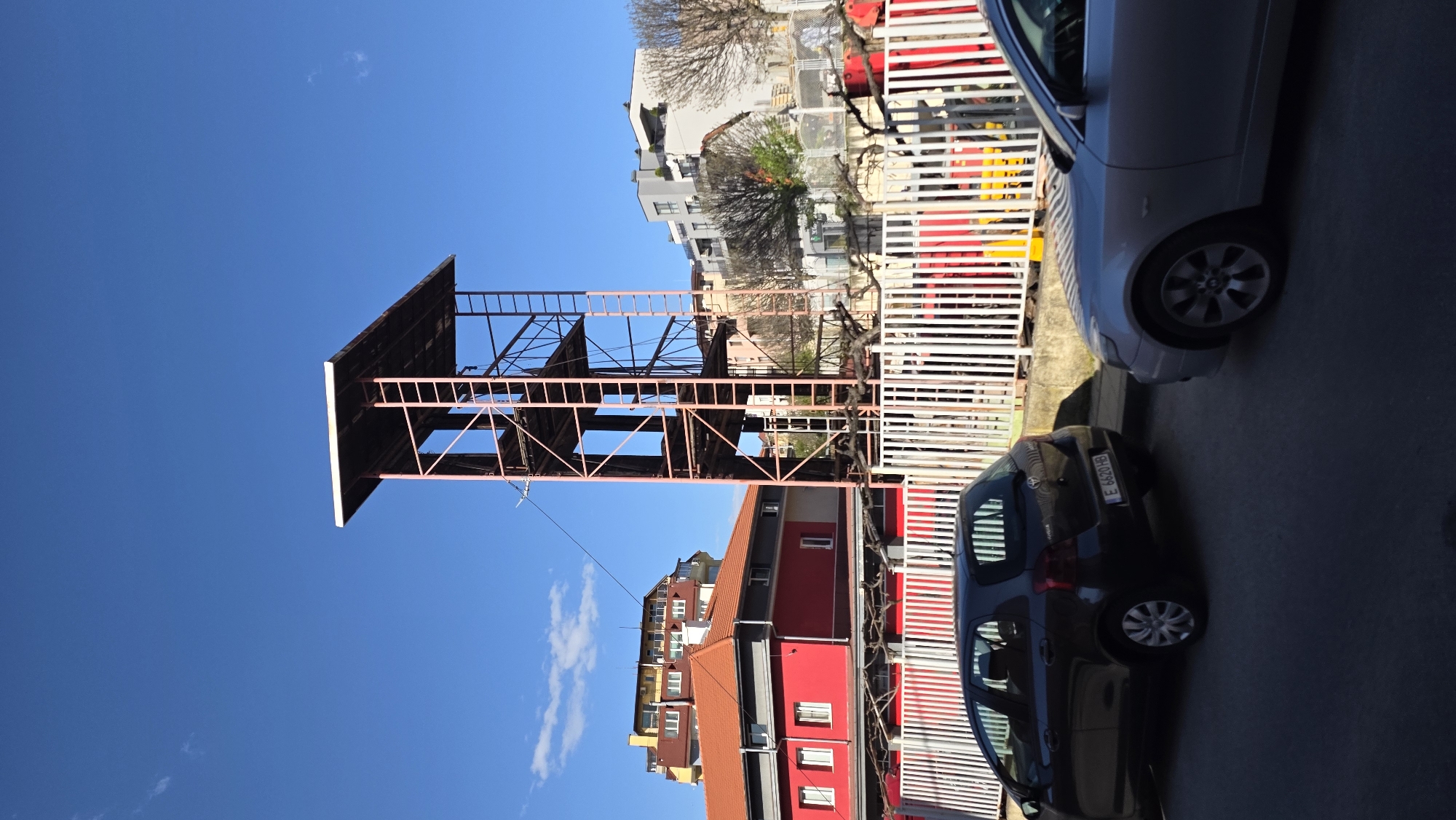 The image depicts a tall, open-frame metal structure under a clear blue sky in Blagoevgrad, Bulgaria. This structure is positioned near a variety of buildings, including multi-story apartments with red and orange roofs. In the foreground, a white fence with red and yellow markings runs vertically. Adjacent to the fence are two parked cars: a silver sedan and a dark-colored hatchback with the license plate "E 6820 HB."